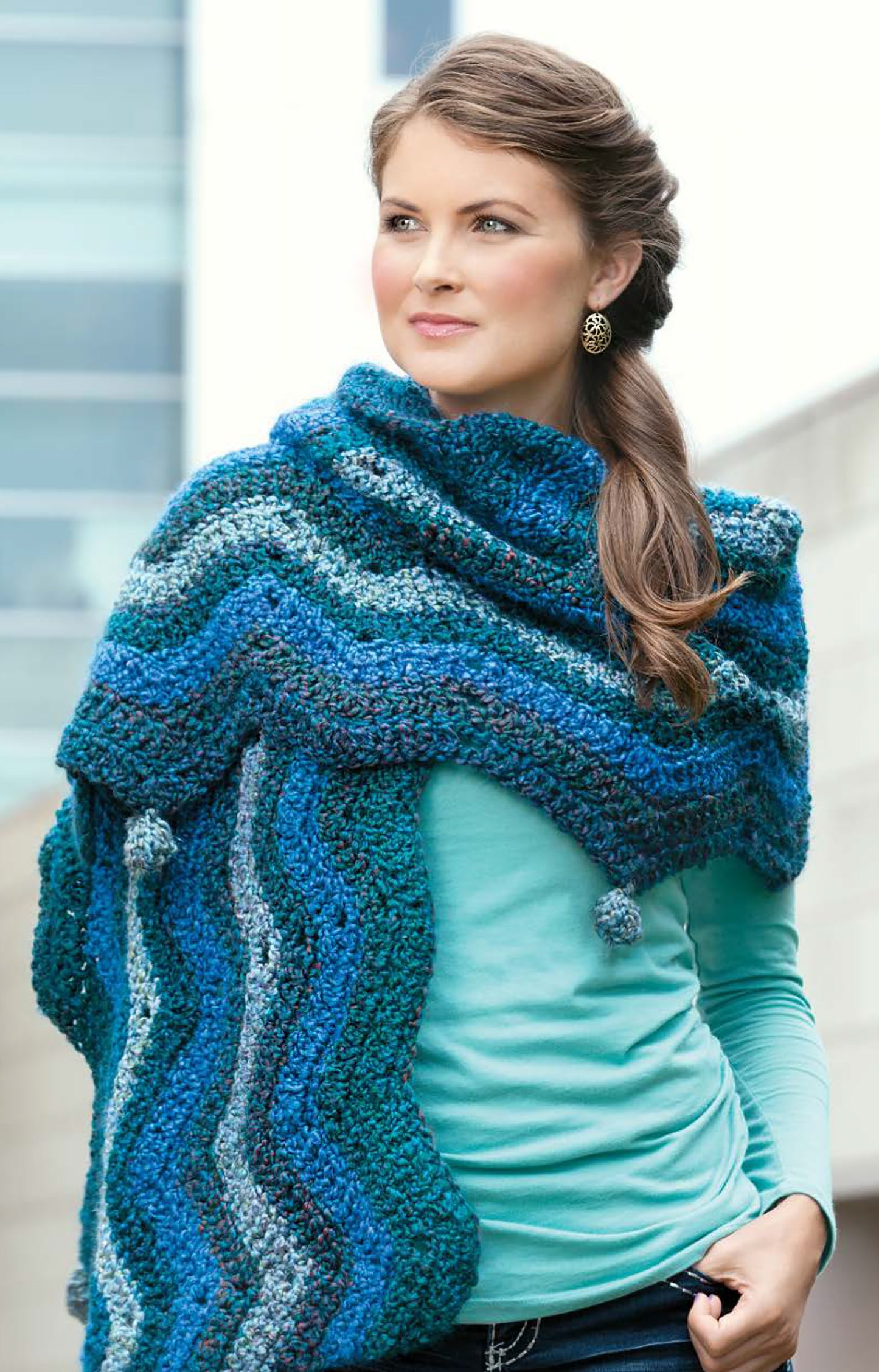 A woman in a bright blue long-sleeve top and the "Cozy Ripples for You & Your Home" poncho by Leisure Arts stands outdoors, gazing to the side with a soft smile. Her brown hair is styled in a loose braid.