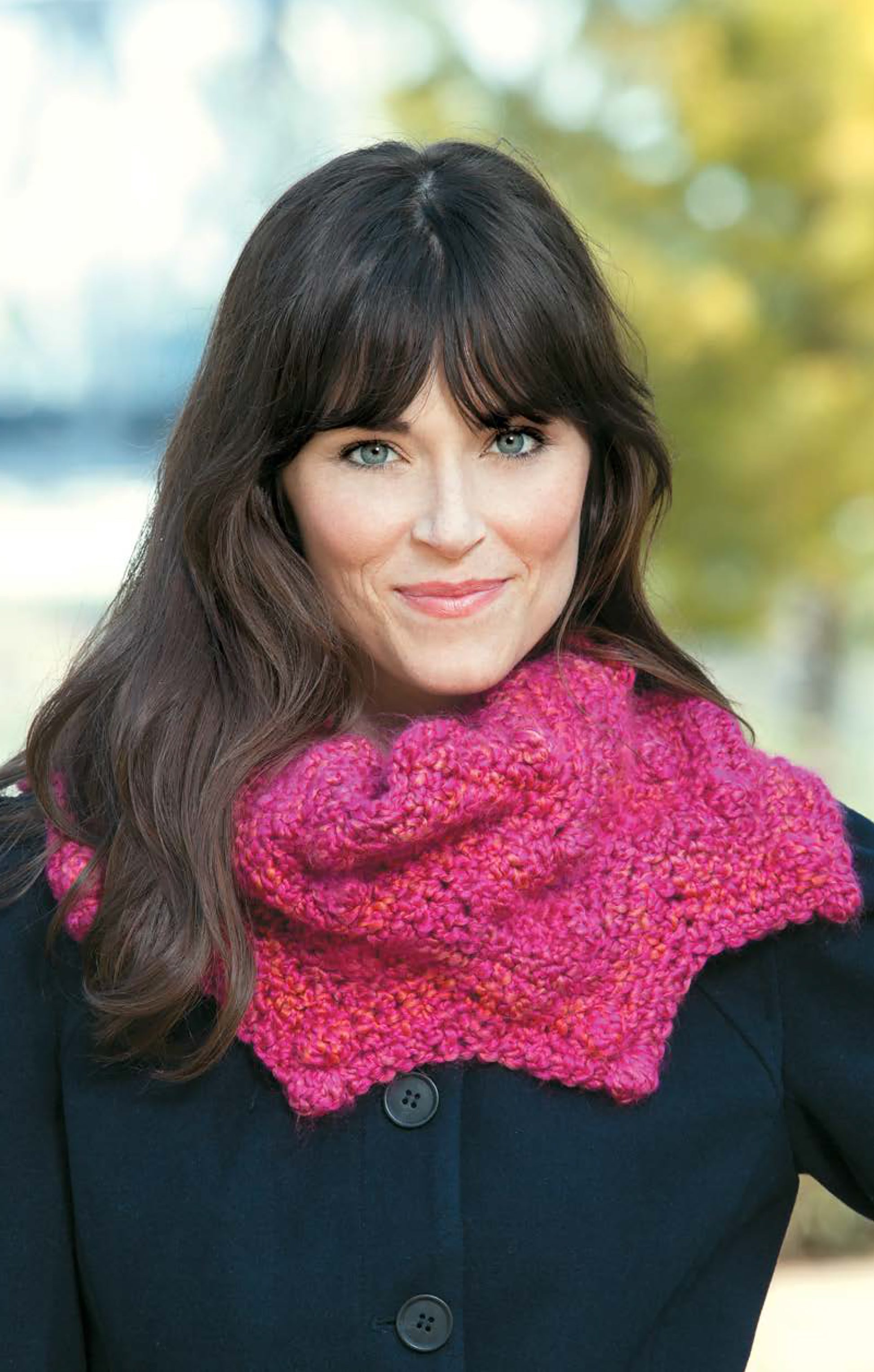 A woman with long brown hair, in a dark coat and the bright pink Cozy Ripples for You & Your Home shawl by Leisure Arts, stands outdoors smiling softly amid blurred greenery and sunlight.