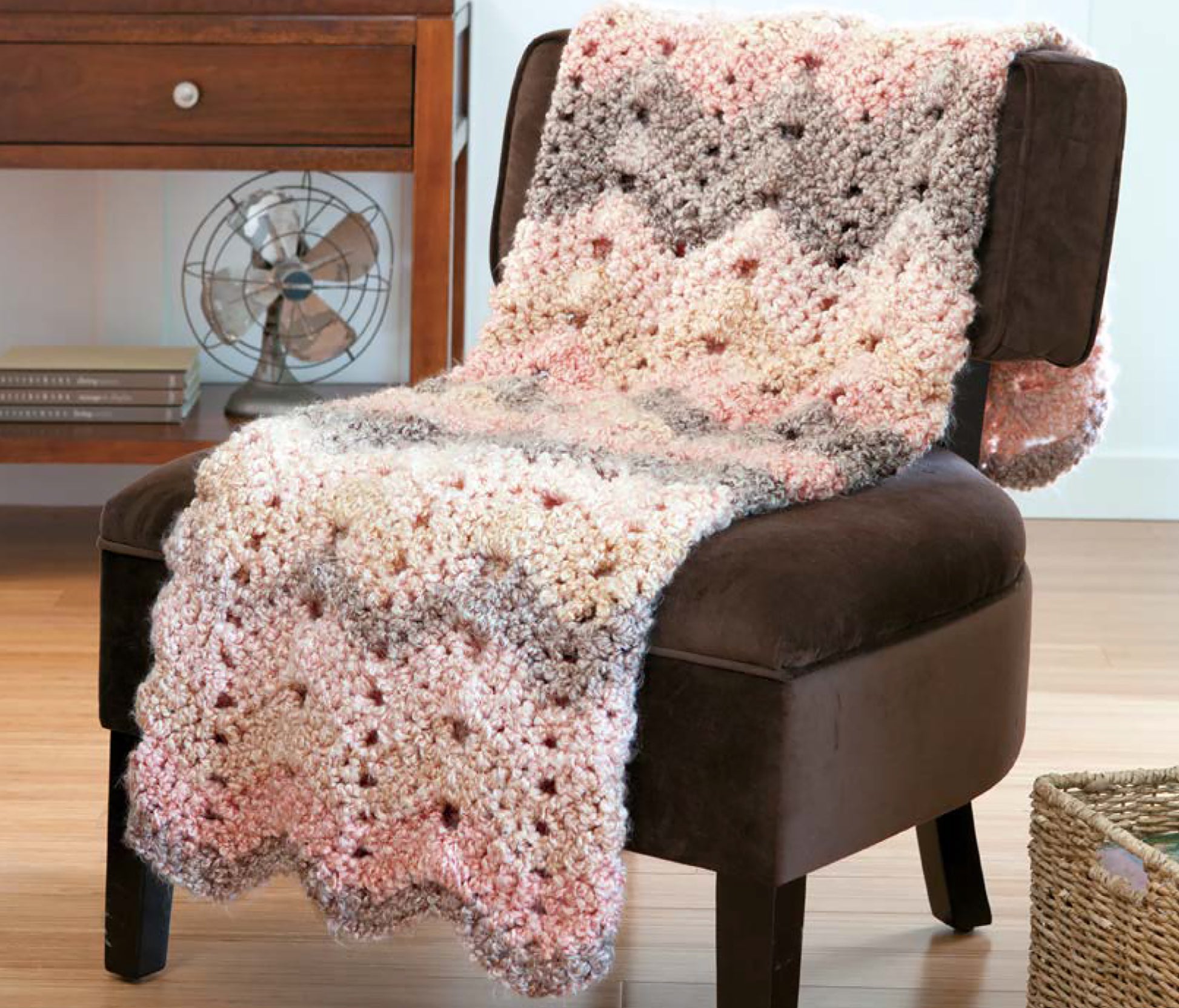 The Cozy Ripples for You & Your Home afghan by Leisure Arts, in pink, cream, and gray, is draped over a dark brown chair in a cozy room with wooden floors, a side table, and a vintage-style fan.