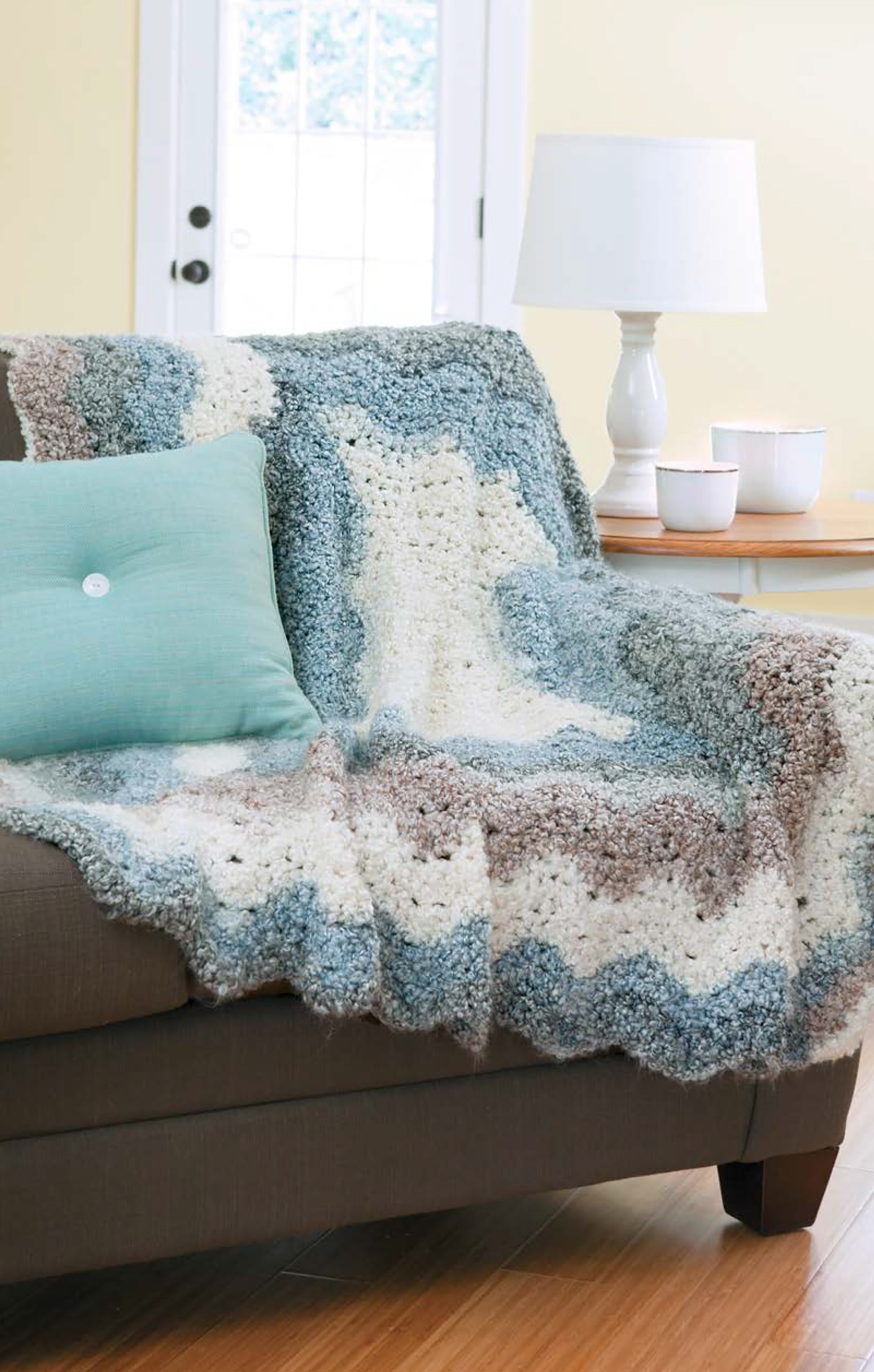 The Cozy Ripples for You & Your Home afghan by Leisure Arts, featuring blue, gray, and cream stripes, is draped over a brown couch beside a light blue pillow. A side table holds a white lamp and two cups near the windowed door.