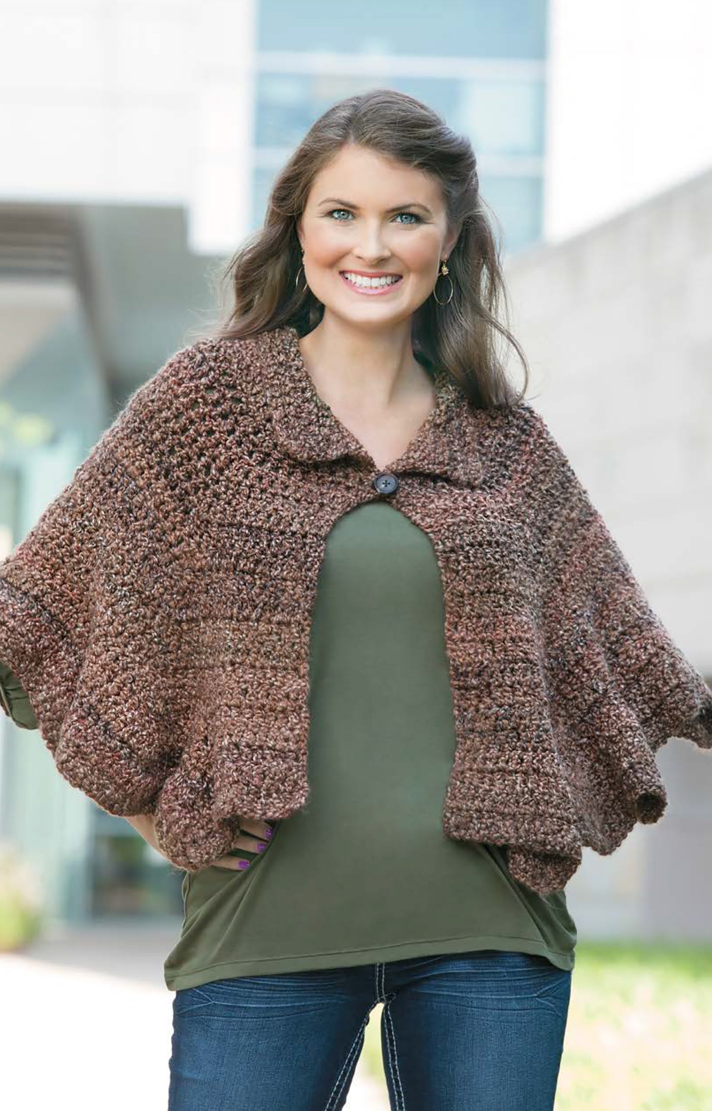 A smiling woman stands outside in blue jeans and an olive shirt, wearing the Cozy Ripples for You & Your Home shawl-capelet by Leisure Arts. The chunky brown crochet wrap features a buttoned collar, set against a backdrop of greenery and a modern building.