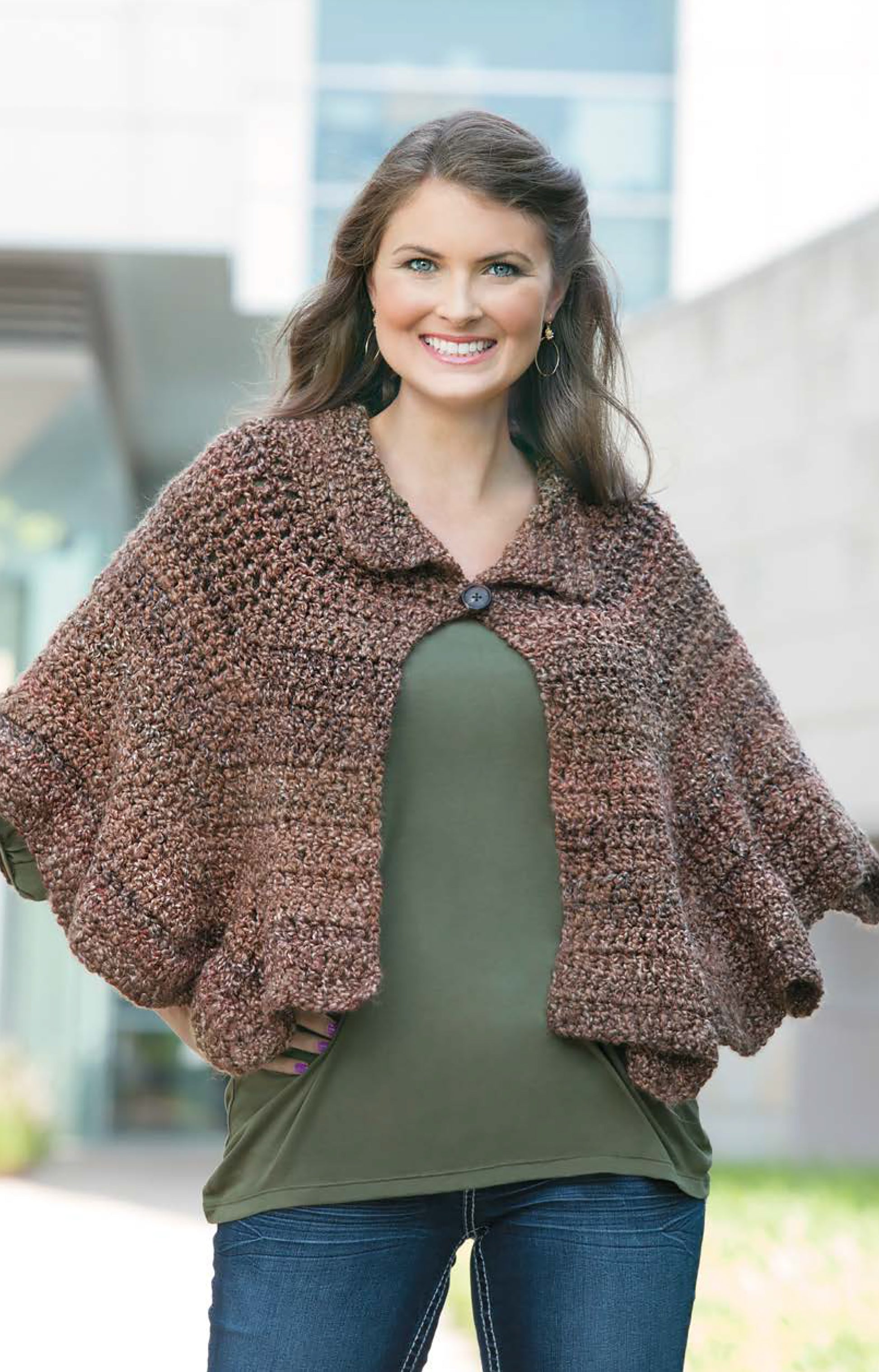 A smiling woman stands outside in blue jeans and an olive shirt, wearing the Cozy Ripples for You & Your Home shawl-capelet by Leisure Arts. The chunky brown crochet wrap features a buttoned collar, set against a backdrop of greenery and a modern building.