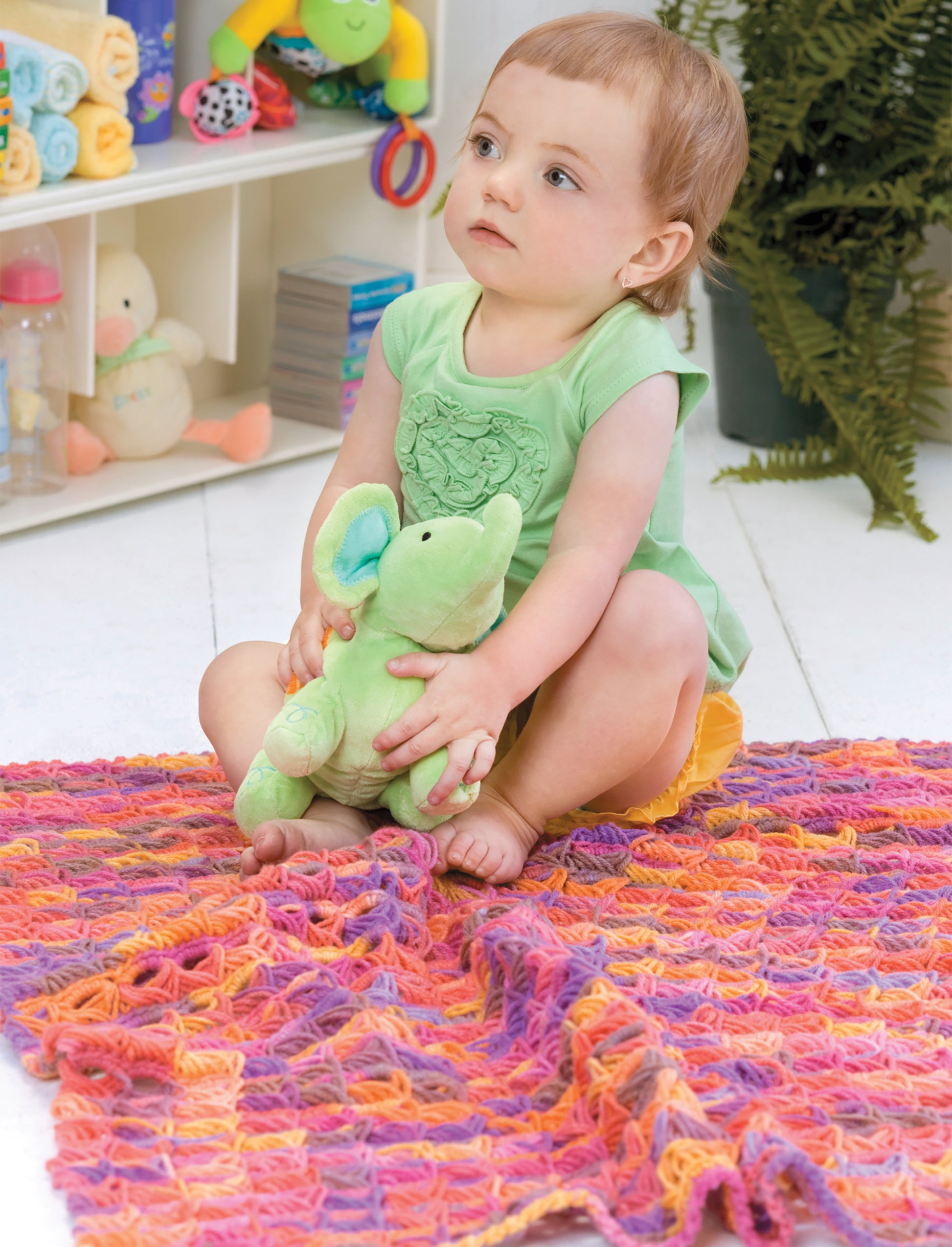 A toddler in green sits on a vibrant pink and orange blanket made with Leisure Arts’ Crochet Bright Baby Blankets Digital Download, holding a green elephant plush. The background features toys, a plant, and neatly organized baby items on shelves.