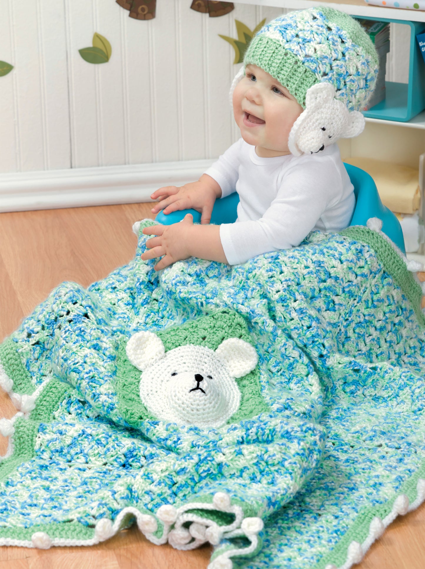 A smiling baby in a white shirt sits on a blue seat, wrapped in the Crochet Bright Baby Blankets by Leisure Arts—a cute, beginner-friendly crochet pattern featuring bear-face details and a matching hat with bear ears. Digital download available.