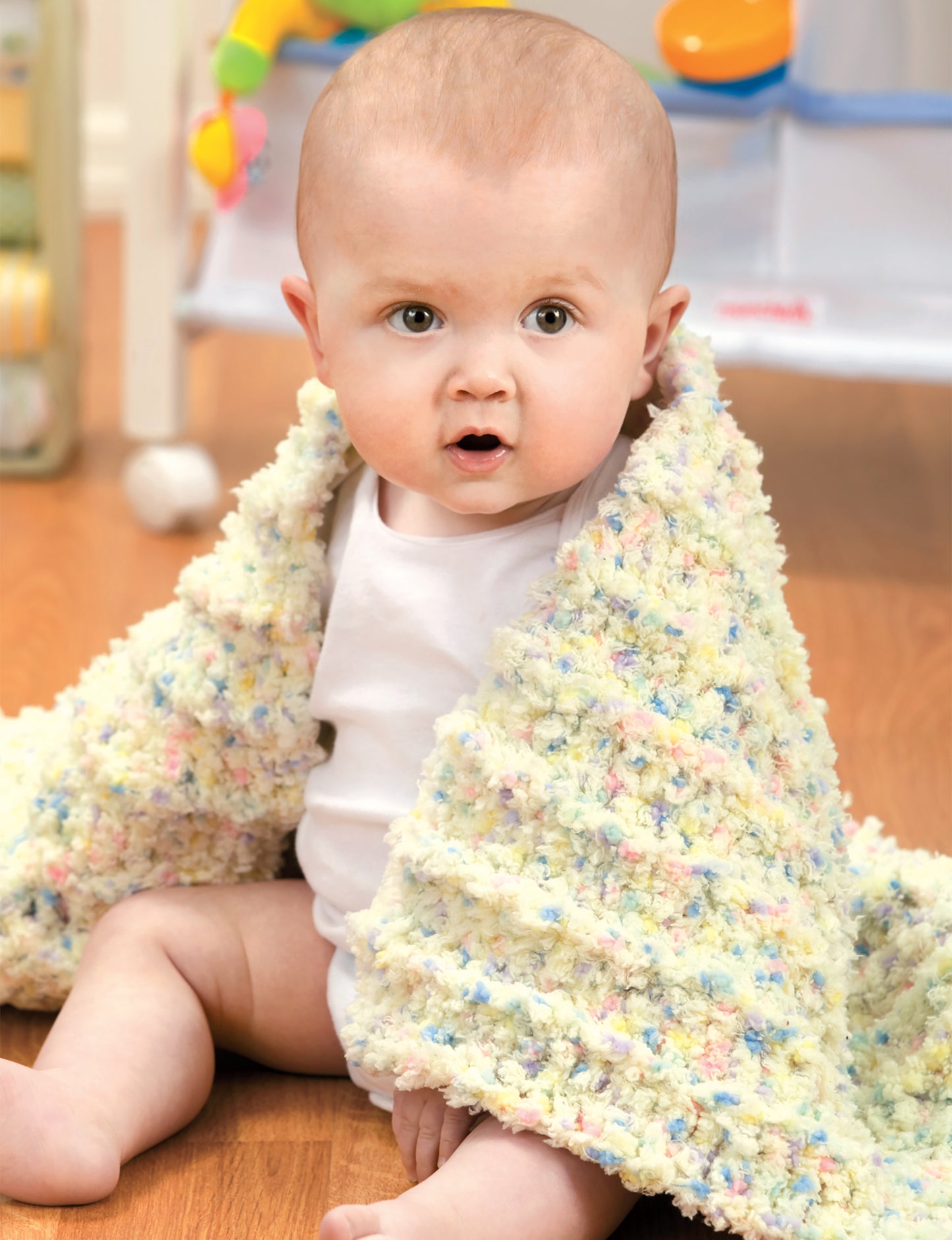 A baby in a white onesie sits on a wooden floor, wrapped in a Leisure Arts Crochet Bright Baby Blanket (Digital Download), featuring cheerful patterns. Toys and baby furniture can be seen in the background as the baby looks surprised.