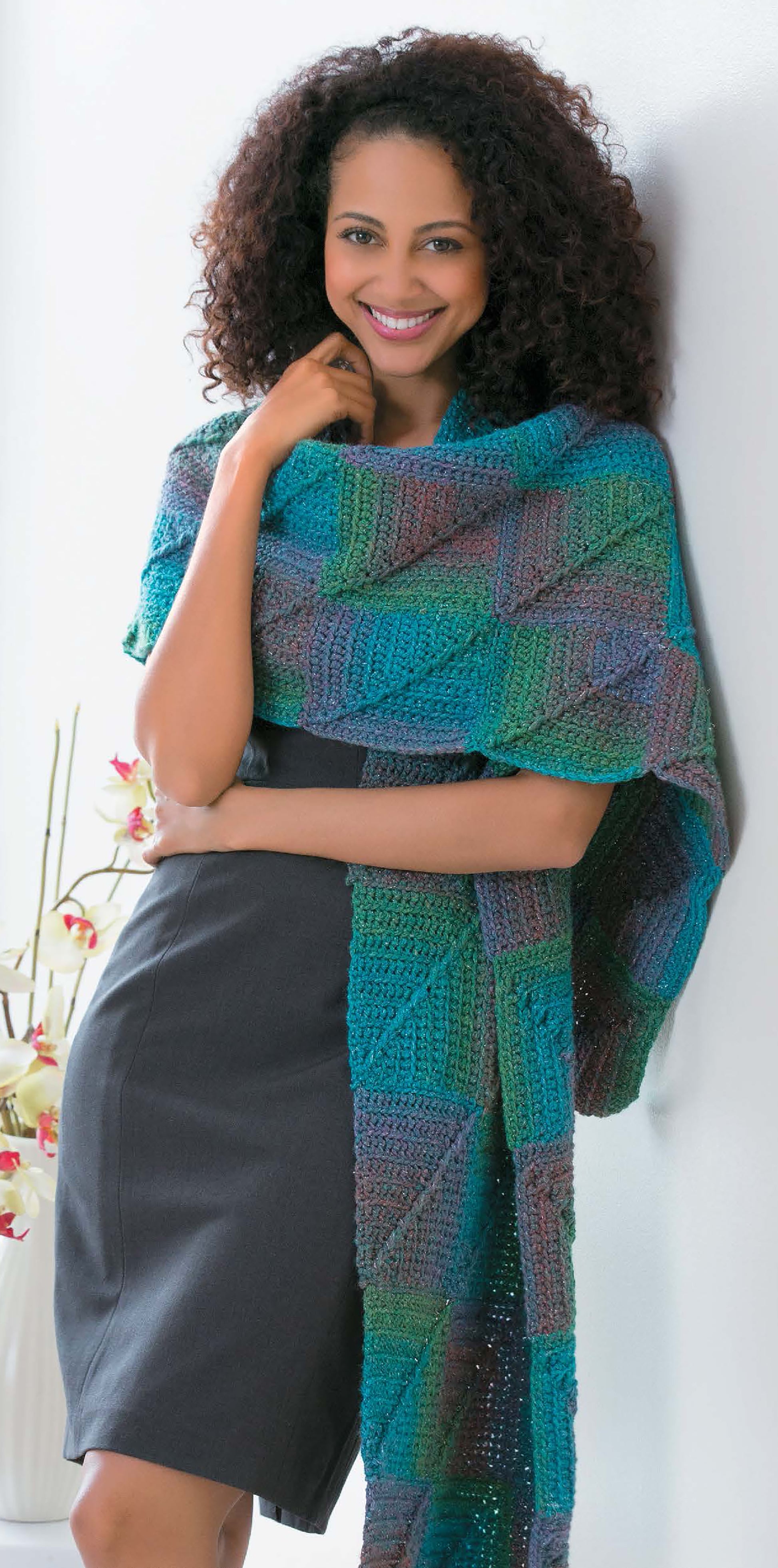 A woman wears a dark dress and a colorful blue, green, and purple shawl from Leisure Arts' Crochet Fashion Accessories, posing by a white wall with one hand on her shoulder and flowers behind her.