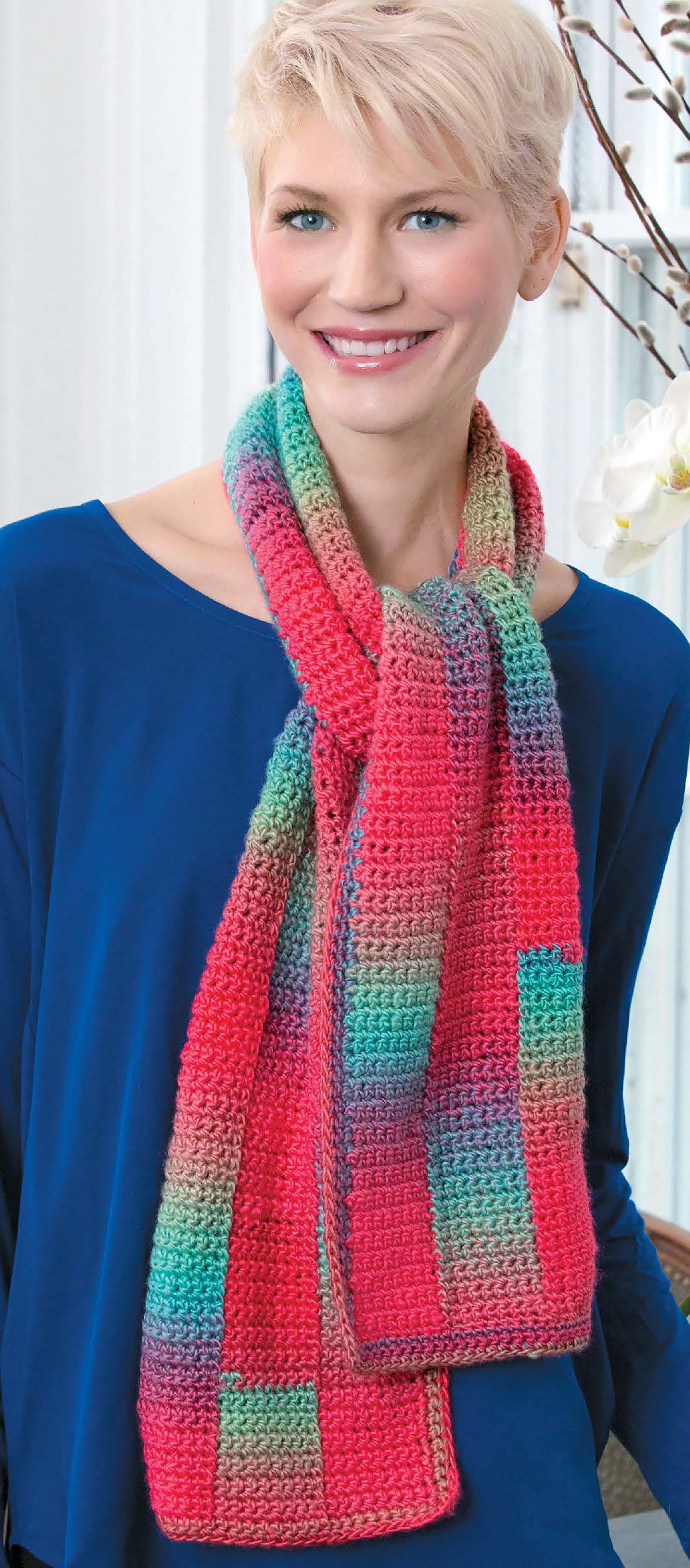 A smiling person with short blonde hair wears a blue top and a vibrant, multicolored scarf made from Crochet Fashion Accessories by Leisure Arts, standing indoors near a window and white flowers.