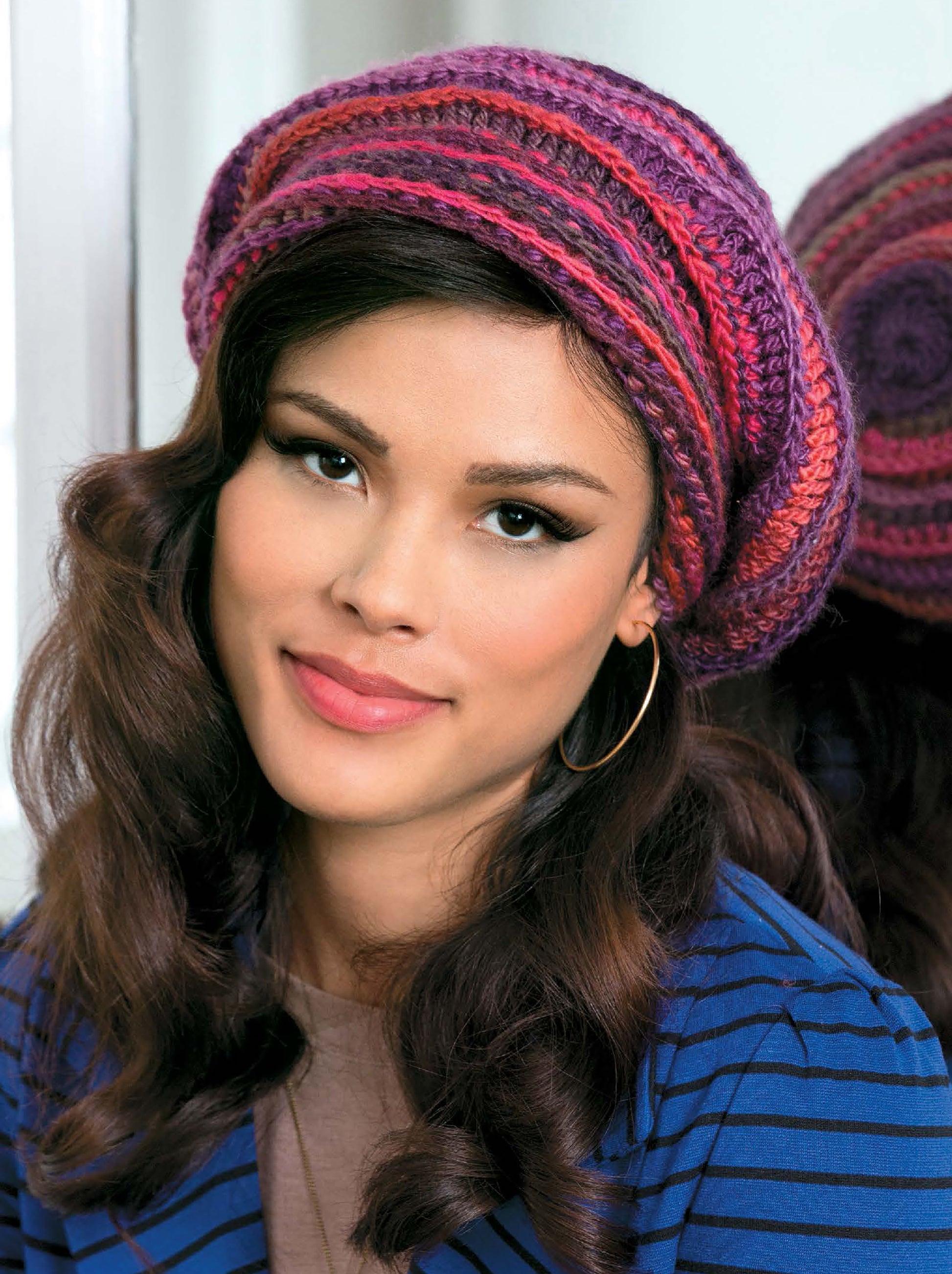 A woman with long, wavy brown hair smiles at the camera, wearing colorful Crochet Fashion Accessories by Leisure Arts—a pink and purple knit beret—paired with a blue striped jacket and large hoop earrings.