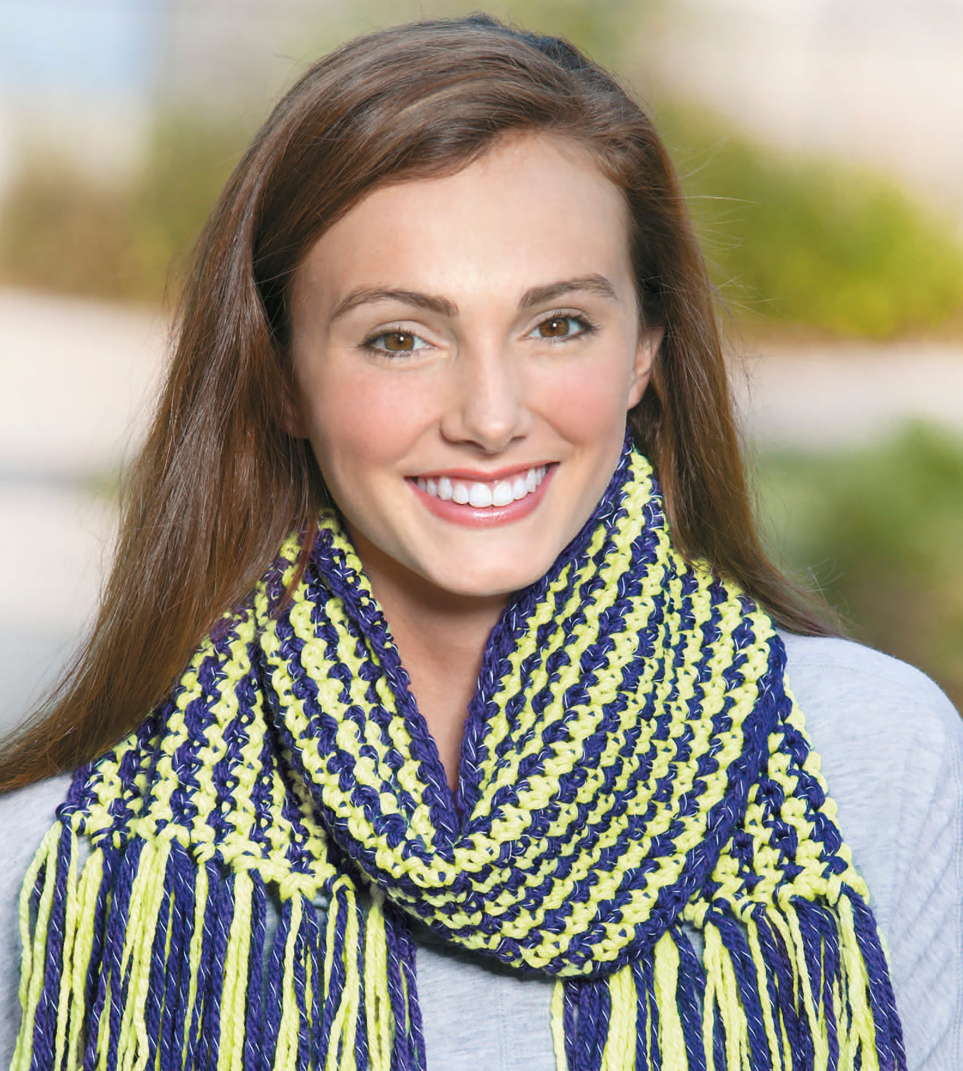 A young woman with long brown hair smiles at the camera, modeling the Light-Reflecting Fashions scarf by Leisure Arts. She wears a blue and yellow striped knit scarf with a light gray top, set against blurred greenery for a cozy seasonal look.