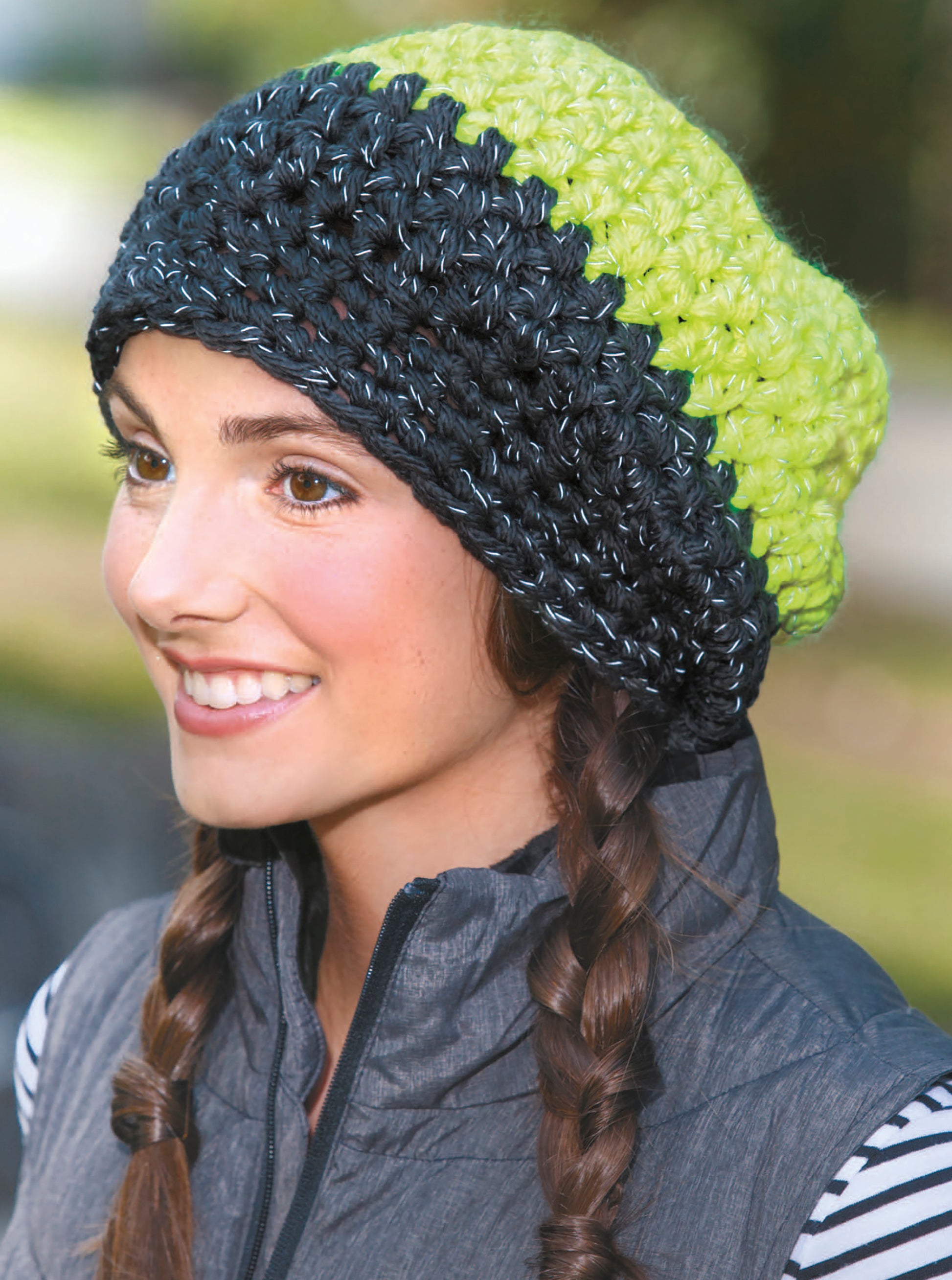 A smiling woman with brown braids models Leisure Arts' Light-Reflecting Fashions—a hand-crocheted black and bright green striped hat—outdoors, where matching scarves and totes are also showcased against a blurred backdrop.