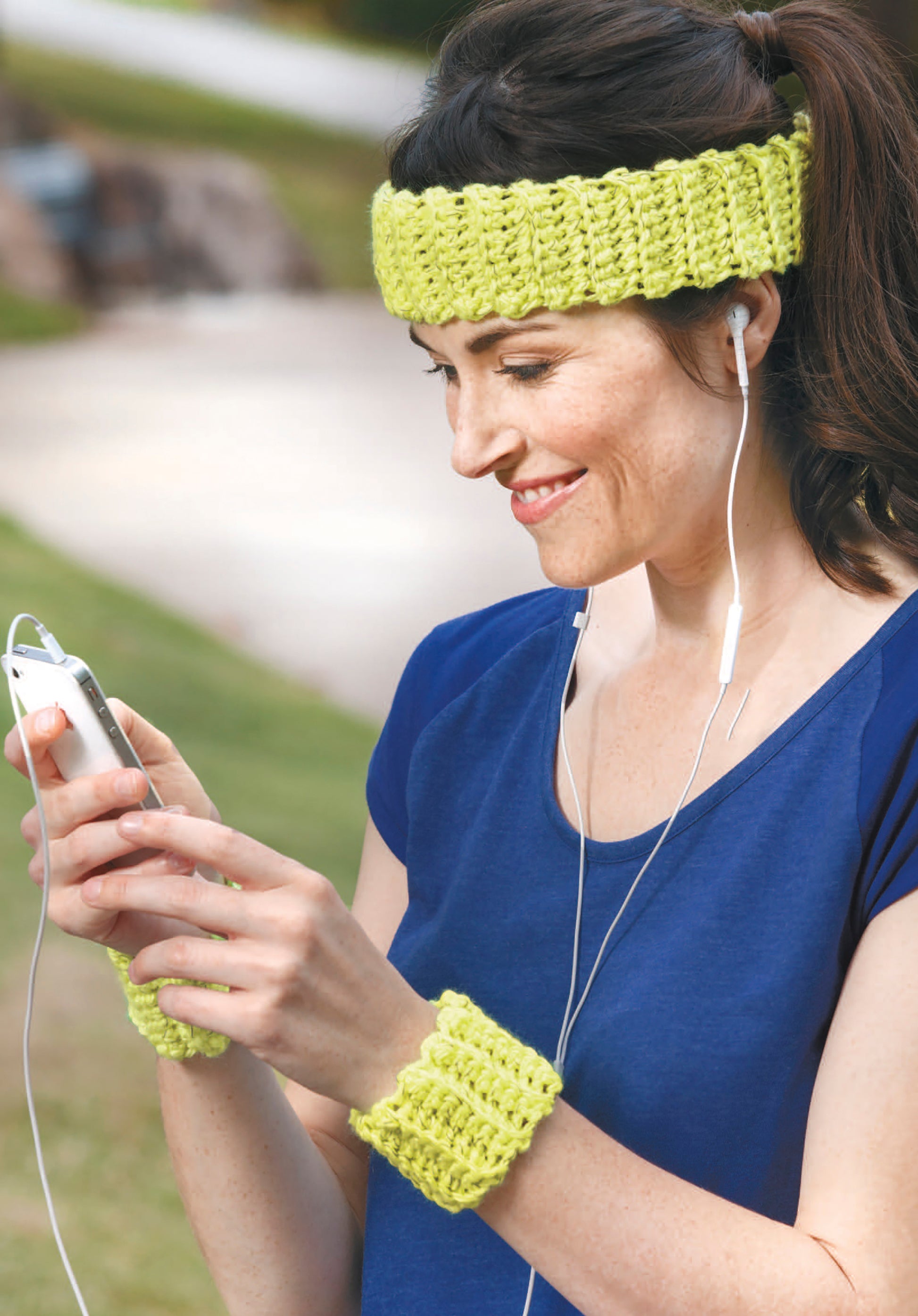 A woman in a blue shirt and yellow headband smiles outdoors, listening to earphones and browsing Leisure Arts’ Light-Reflecting Fashions for unique scarves and dog sweaters on her smartphone.