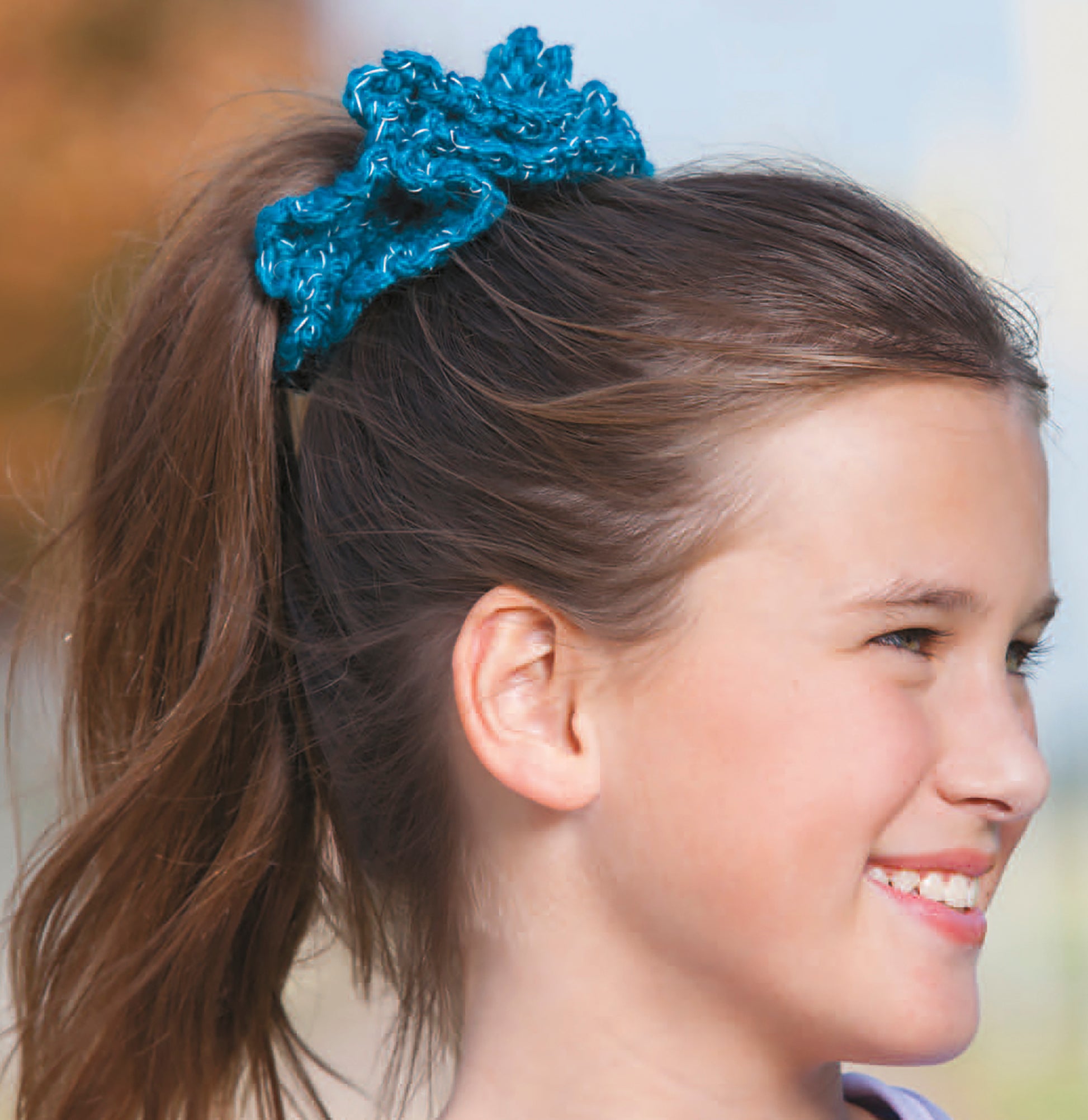 A girl with brown hair in a ponytail, smiling and wearing a bright blue crocheted scrunchie, is shown outdoors in profile view carrying a stylish "Light-Reflecting Fashions" tote from Leisure Arts.