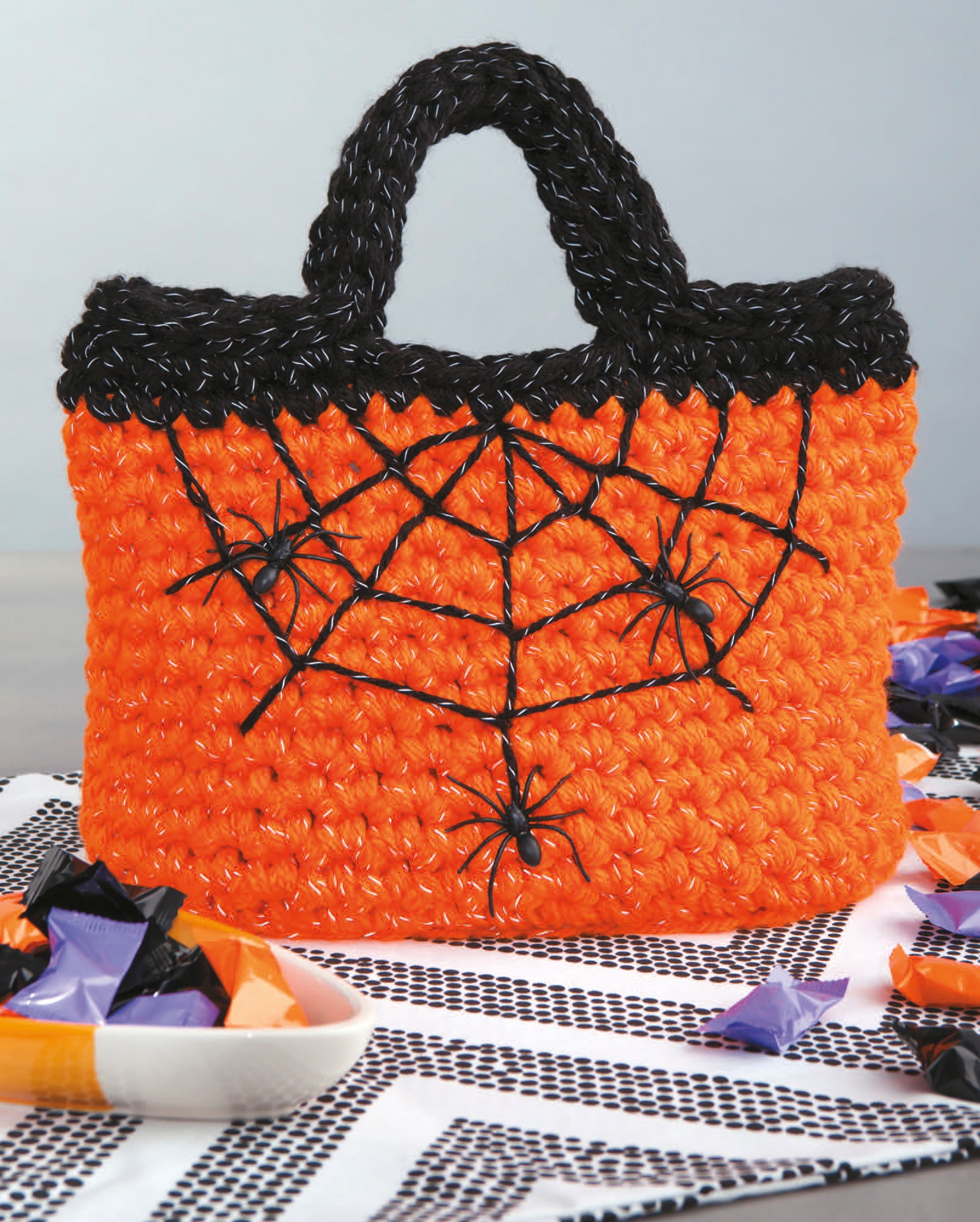 The Light-Reflecting Fashions tote bag by Leisure Arts is orange with black handle and trim, embroidered spiderwebs, and plastic spiders—ideal for Halloween treats or accessorizing. Shown on a black-and-white table runner.