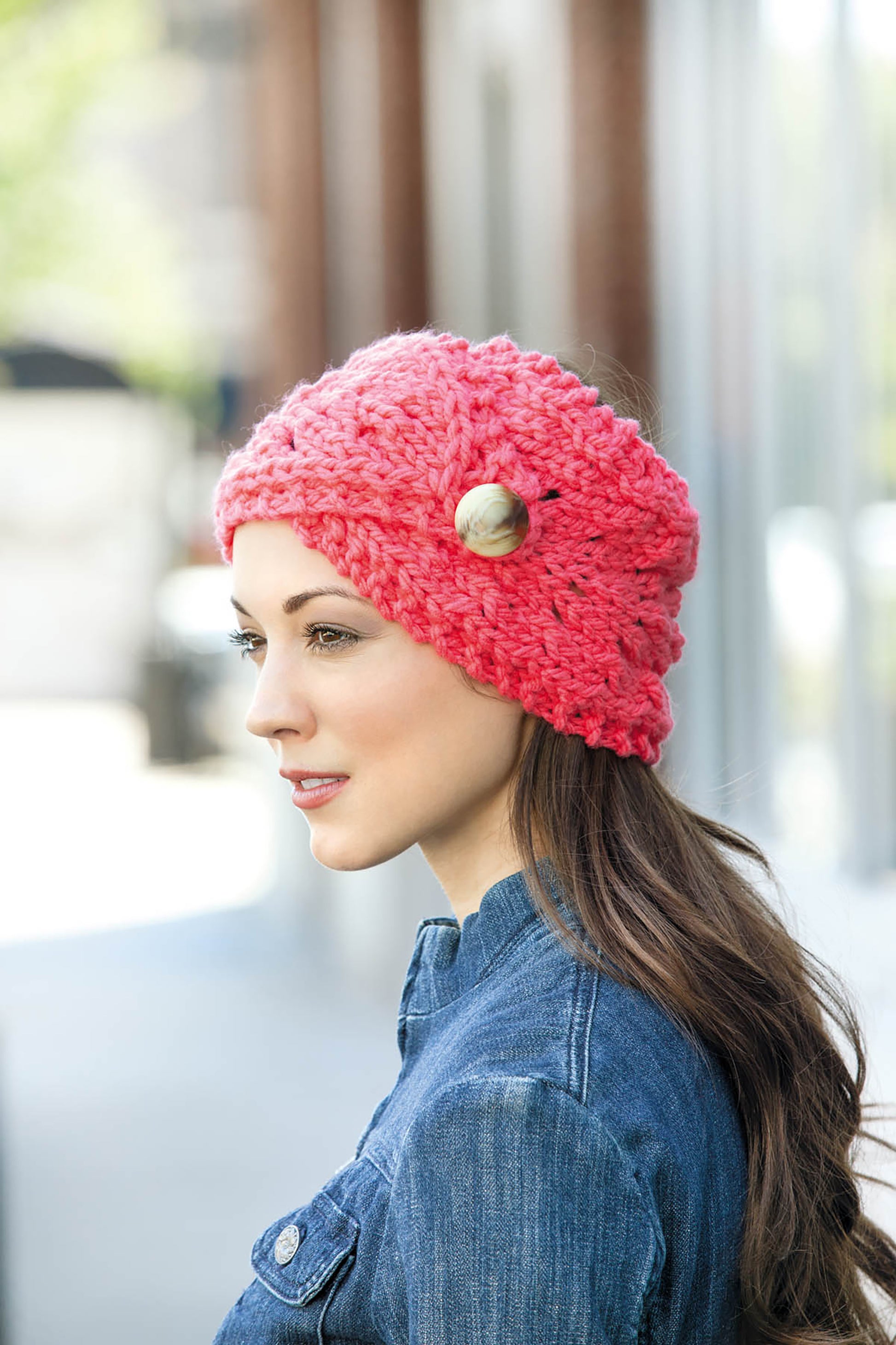 A woman with long brown hair wears a chunky pink knit headband from Cool-Weather Family Fashions by Leisure Arts, featuring a button detail, paired with a denim jacket outdoors in soft daylight.
