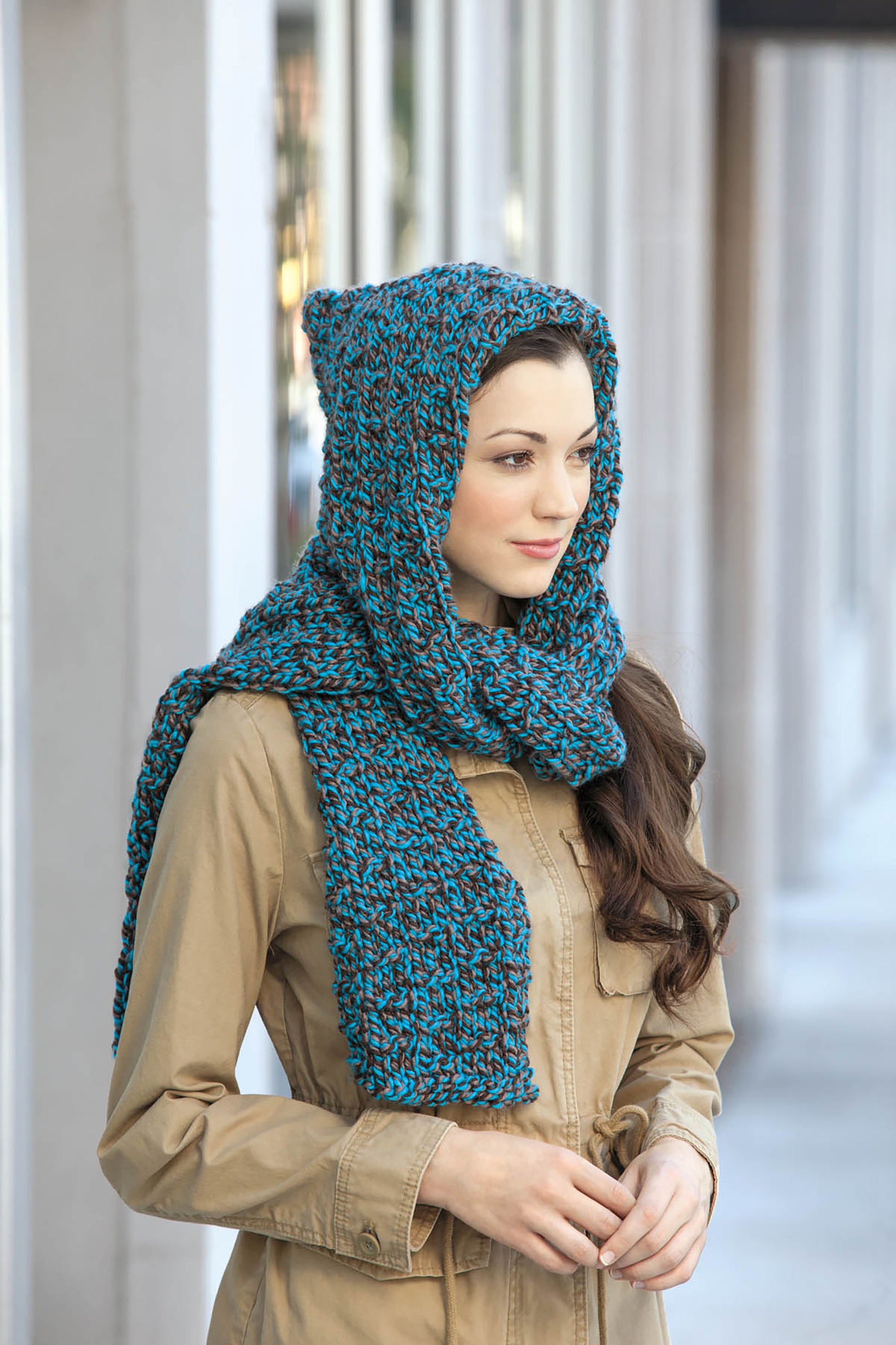 A woman in a tan coat and chunky blue knit hooded scarf from Leisure Arts’ Cool-Weather Family Fashions stands with hands clasped, looking to the side outdoors by a building with tall windows and pillars.