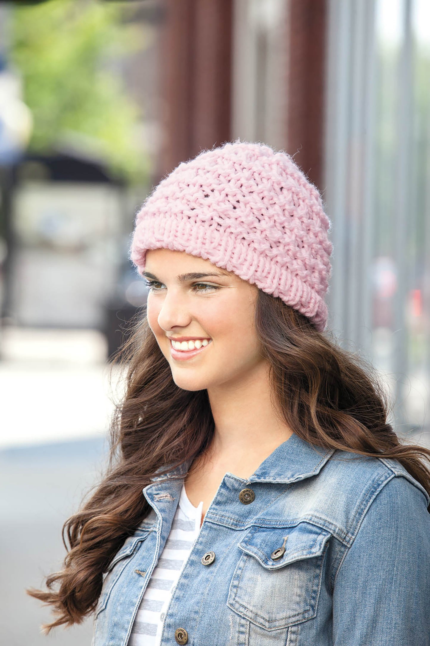 A young woman with long brown hair smiles outdoors on a sunny day, wearing a pink knit beanie from Cool-Weather Family Fashions by Leisure Arts and a denim jacket.