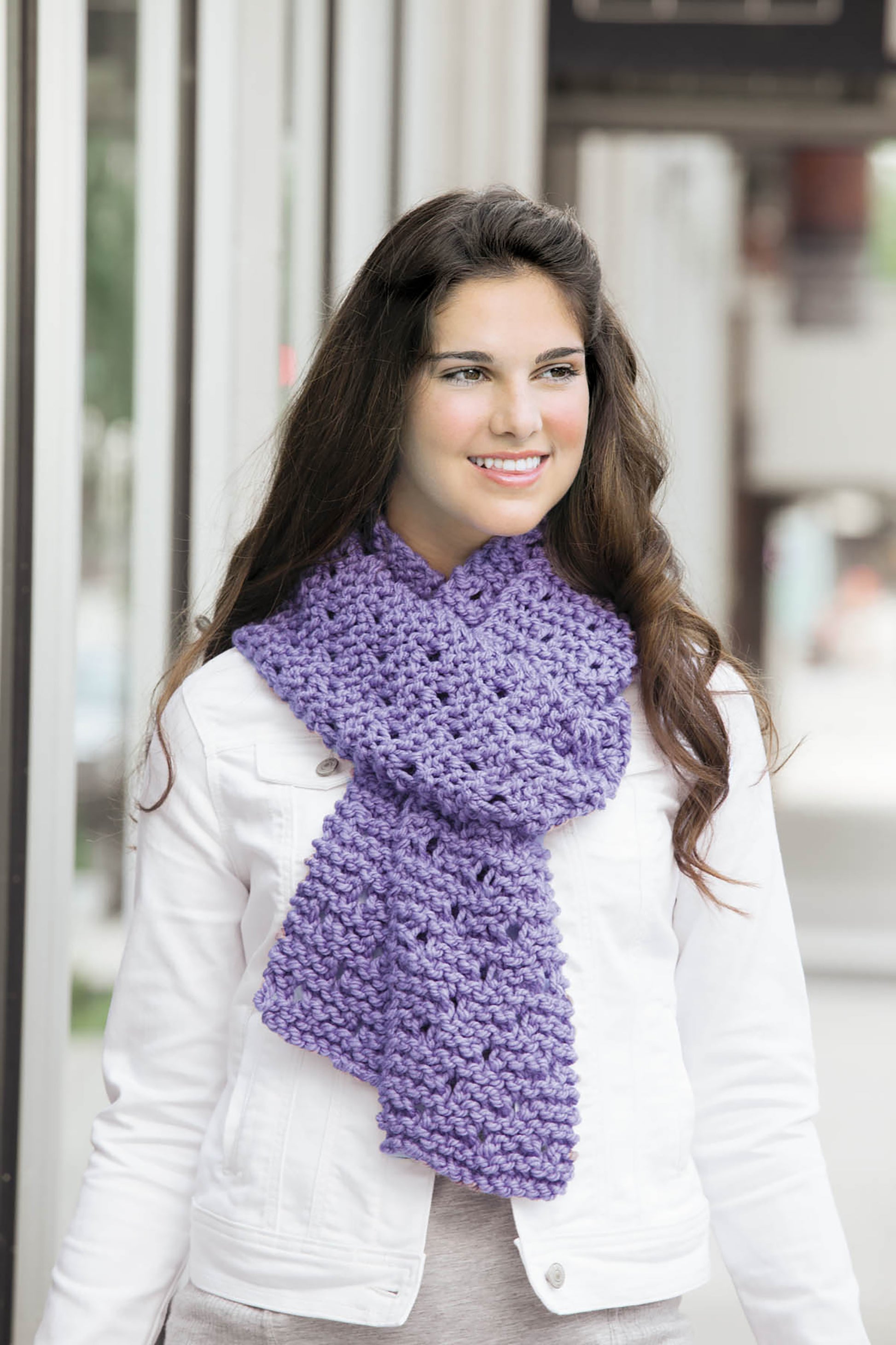 A woman with long brown hair wears a white jacket and a chunky purple scarf made from super bulky yarn, featured in Cool-Weather Family Fashions by Leisure Arts, as she stands outside smiling and looking slightly to the side.
