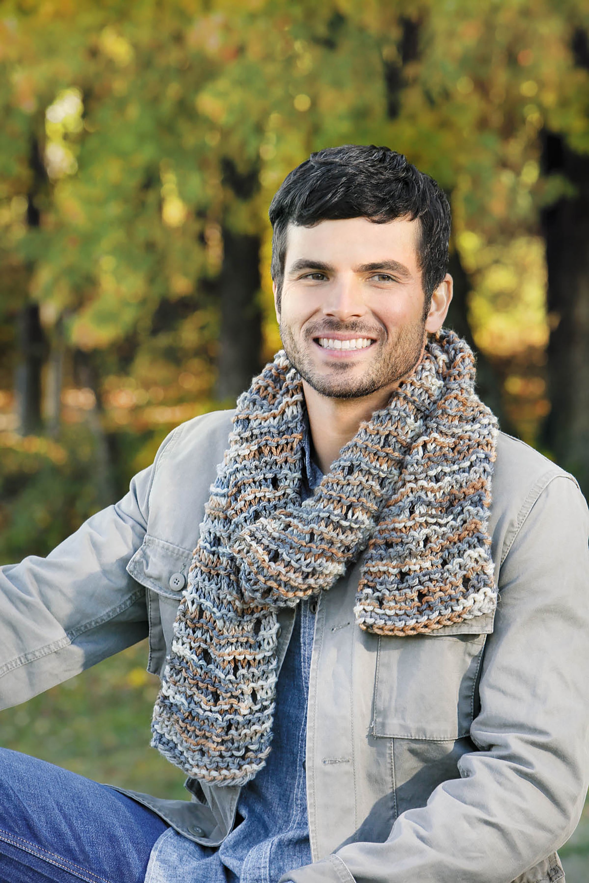 A smiling man with short dark hair and a beard sits outdoors in front of green trees, wearing a grey jacket and a chunky super bulky yarn scarf featured in Cool-Weather Family Fashions by Leisure Arts.