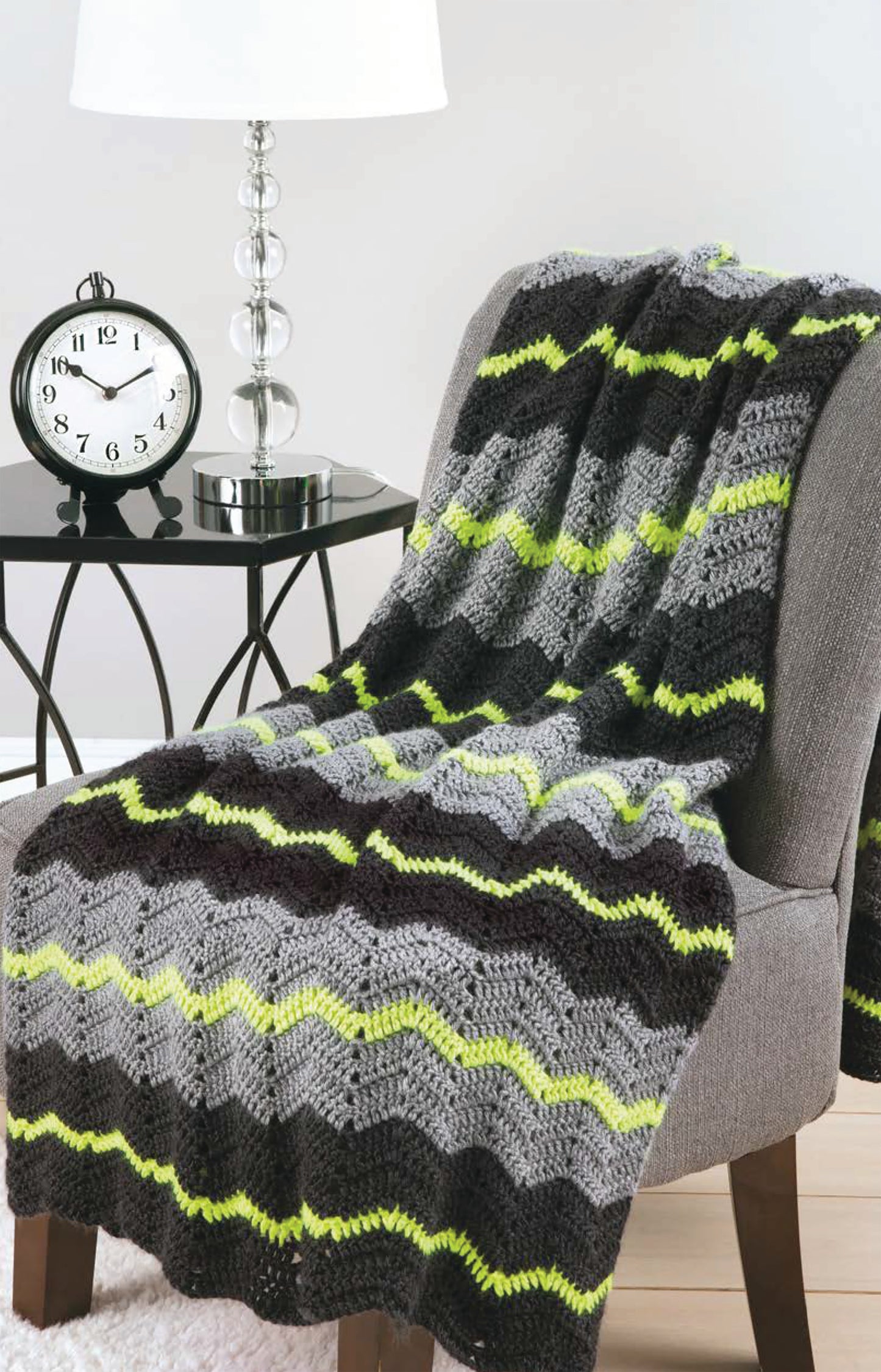 A gray, black, and neon green chevron-pattern throw—made from Chevron Afghans Digital Download by Leisure Arts—is draped over a gray armchair; a black side table with a clock and lamp sits in the background.