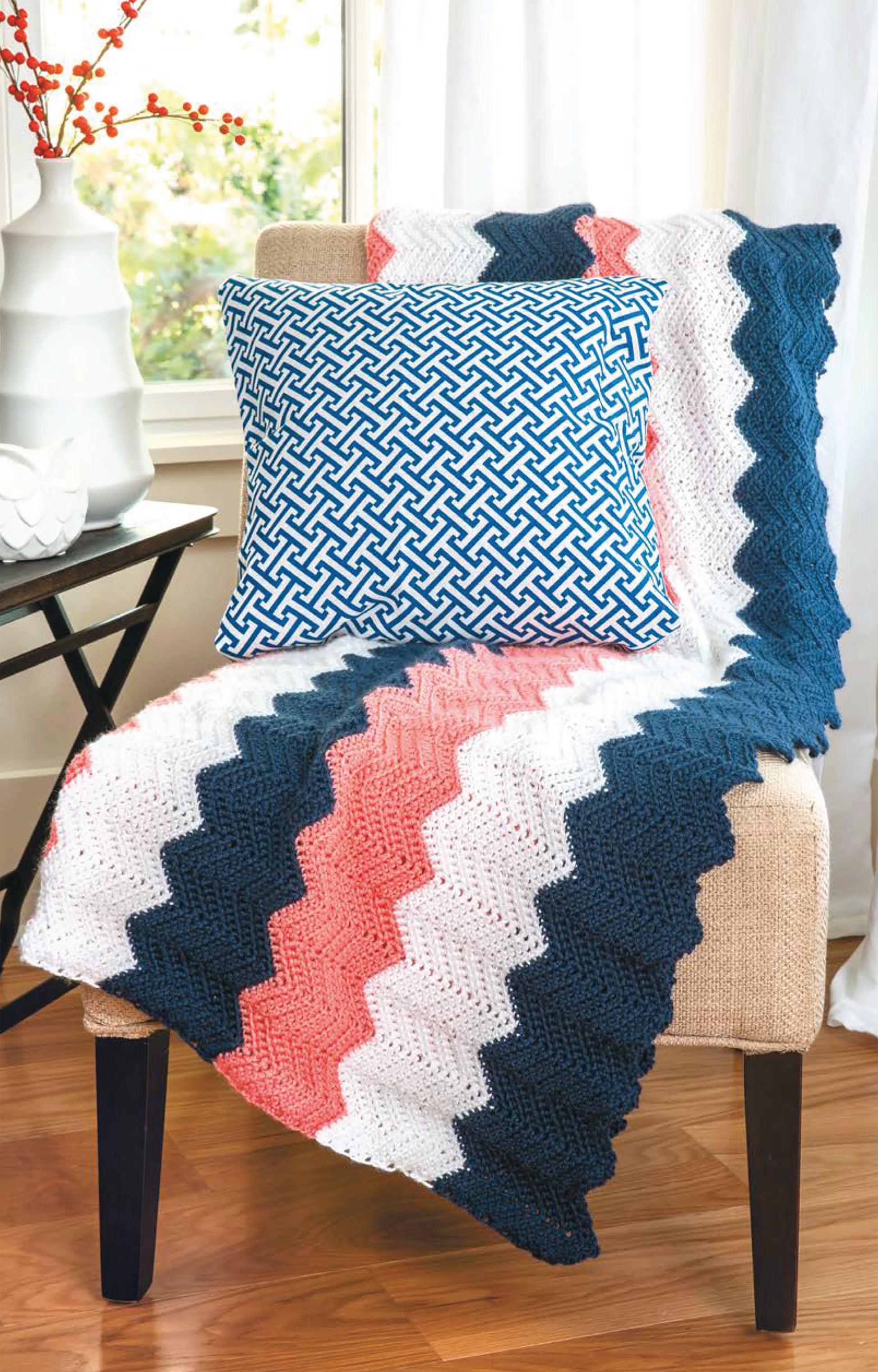 A beige chair showcases a chevron-patterned crochet throw made with Caron Simply Soft yarn from the Chevron Afghans Digital Download by Leisure Arts, topped by a blue and white geometric pillow. A small table and white vase are in the background.