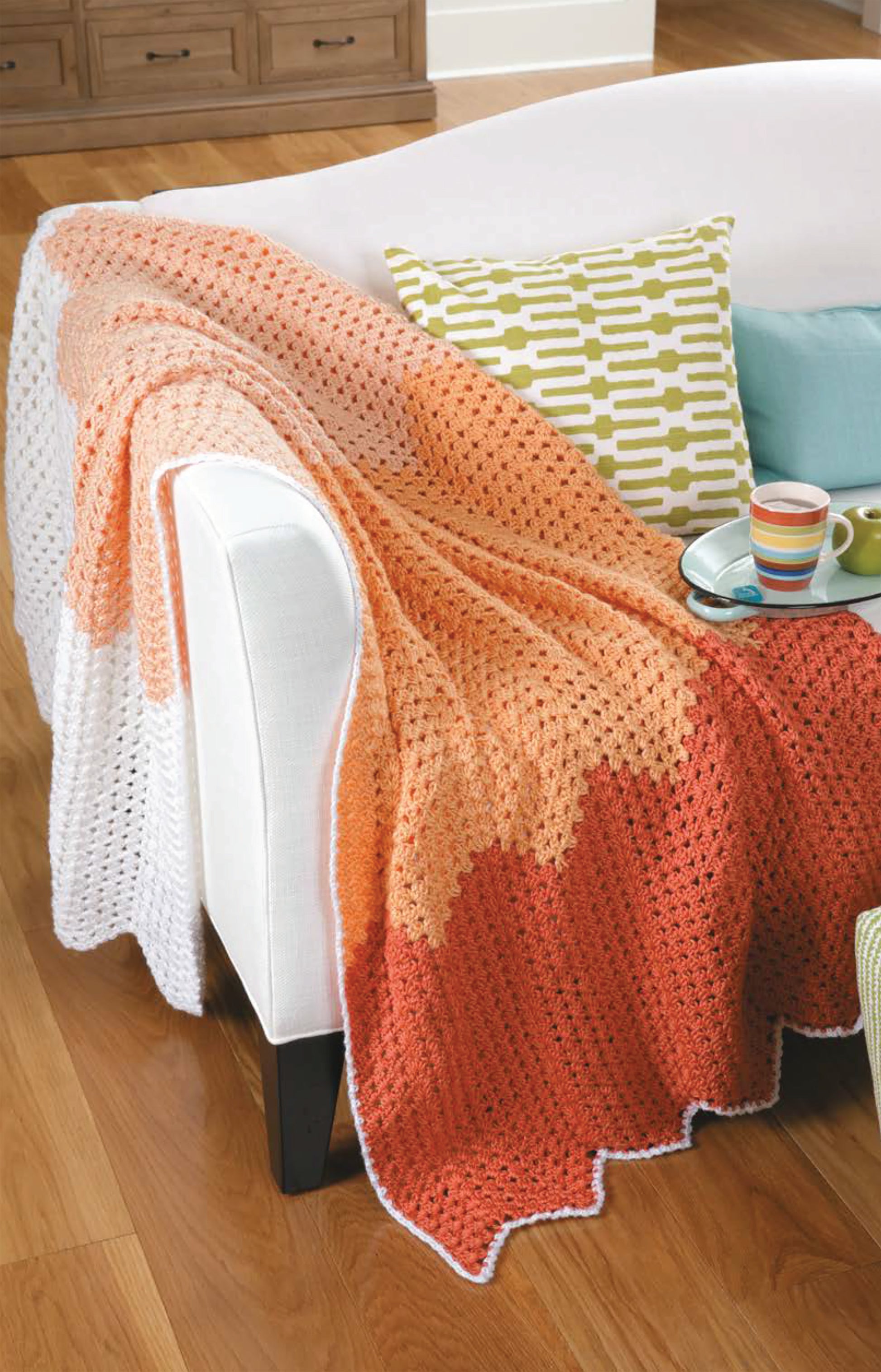 A white sofa with a green and white patterned pillow displays a Chevron Afghans Digital Download blanket by Leisure Arts in white, peach, and orange. A tray with a mug and bowl sits on the wooden floor nearby.