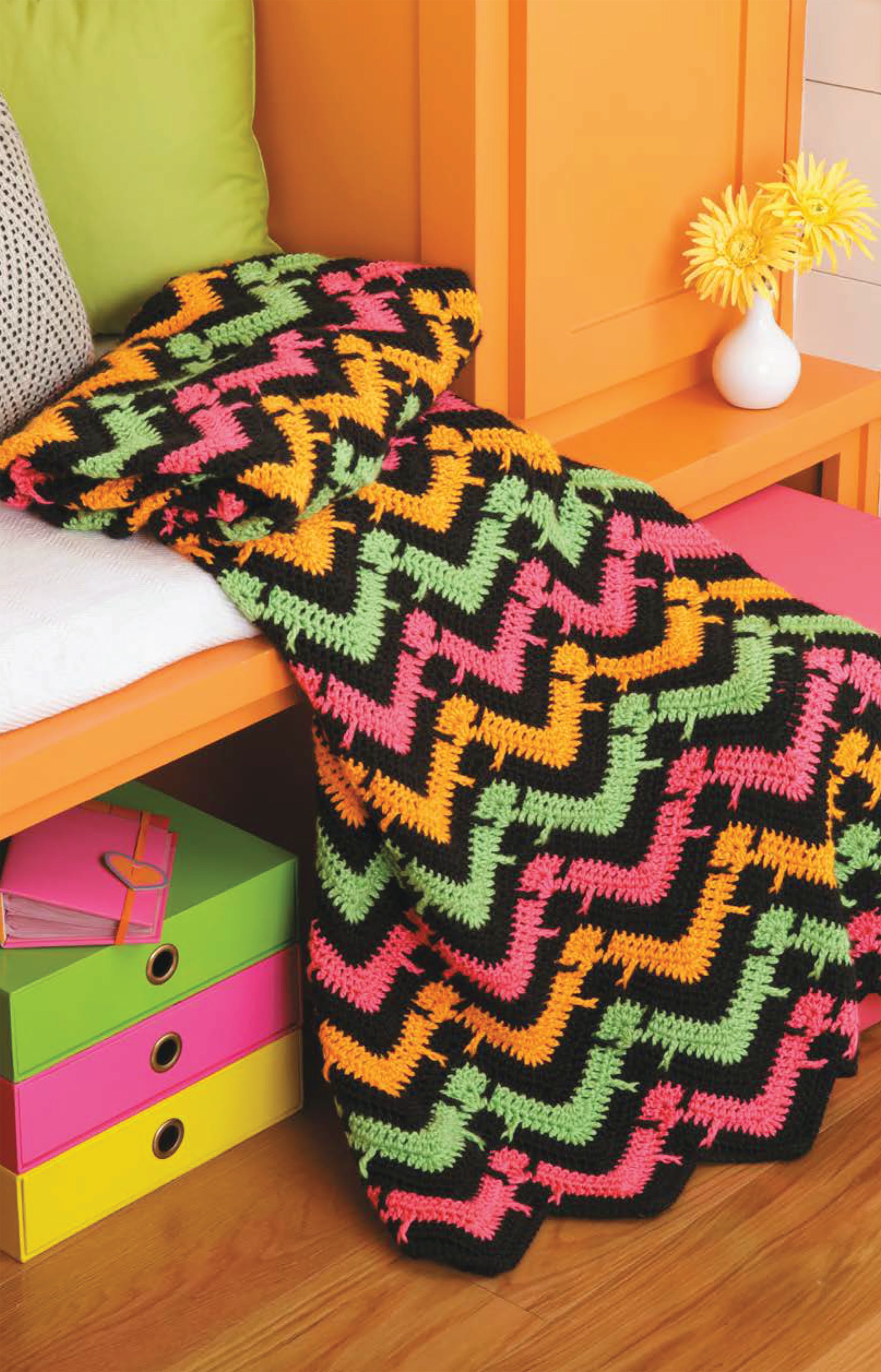 A vibrant blanket from the Chevron Afghans Digital Download by Leisure Arts is draped over an orange bench, beside colorful storage boxes and a vase with a yellow flower.