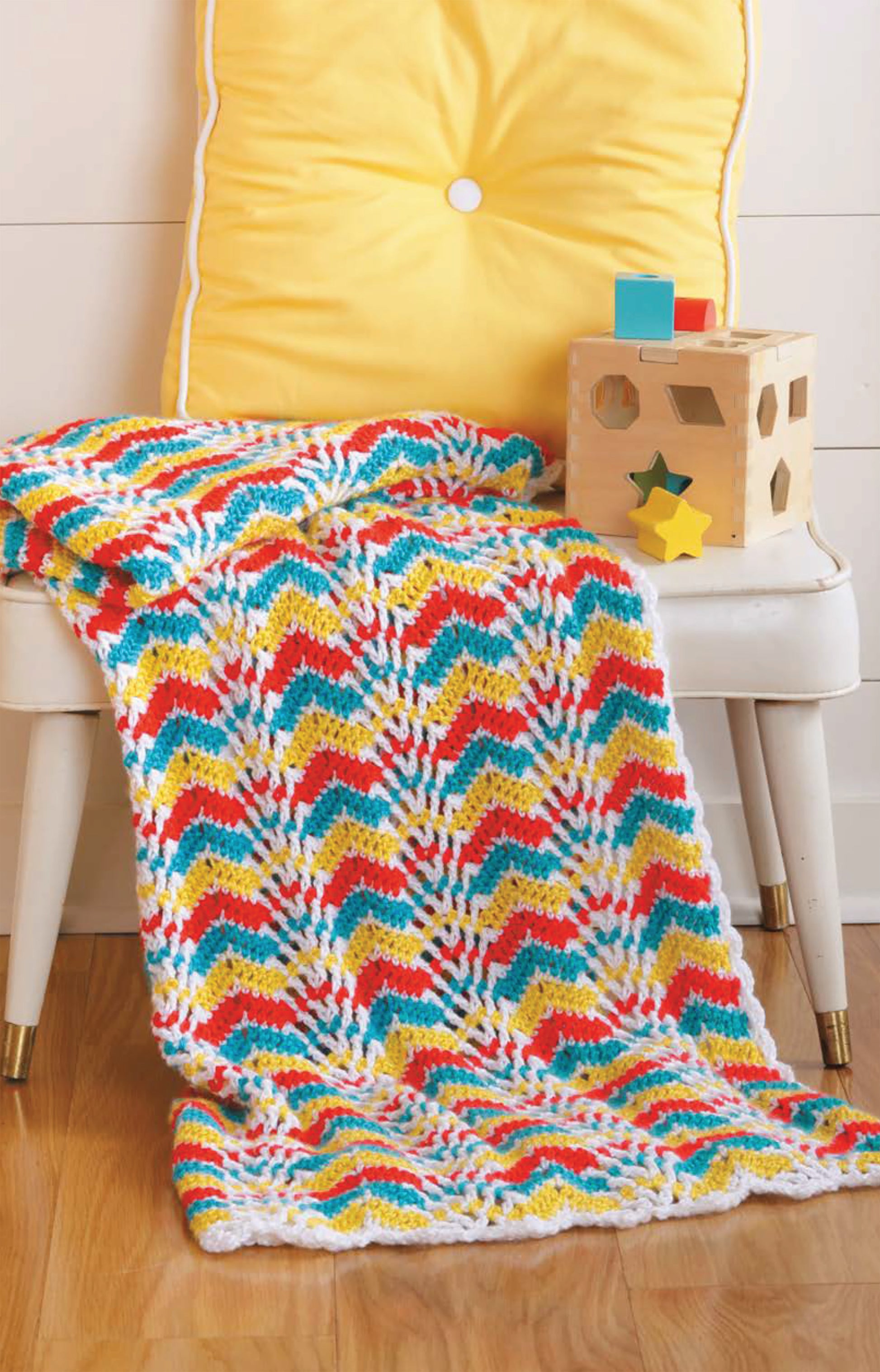 A white chair with a yellow cushion displays a Chevron Afghans Digital Download blanket by Leisure Arts in colorful zigzag patterns. A wooden shape-sorting toy rests on the seat—ideal for crochet throw enthusiasts.