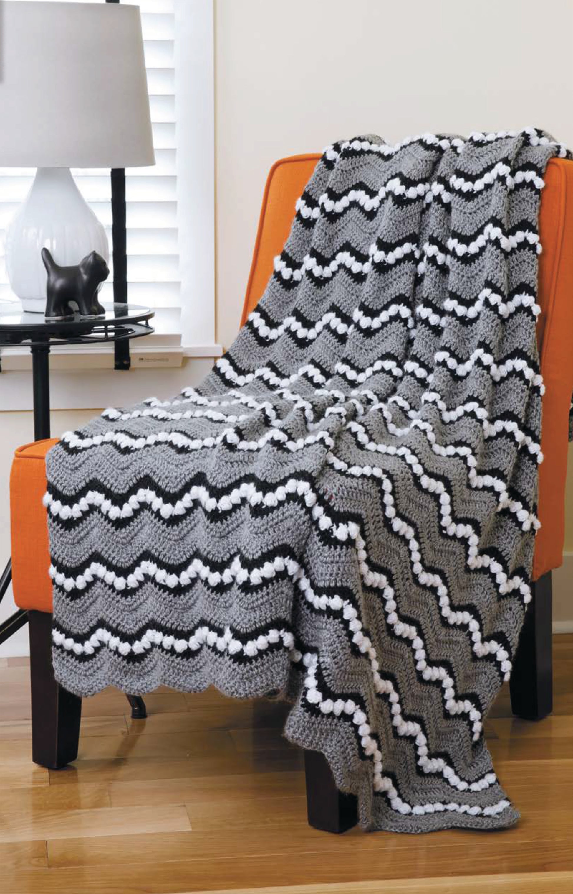 A Chevron Afghans Digital Download from Leisure Arts features gray, black, and white chevron patterns with bobble stitch accents—draped over an orange chair for cozy crochet inspiration.