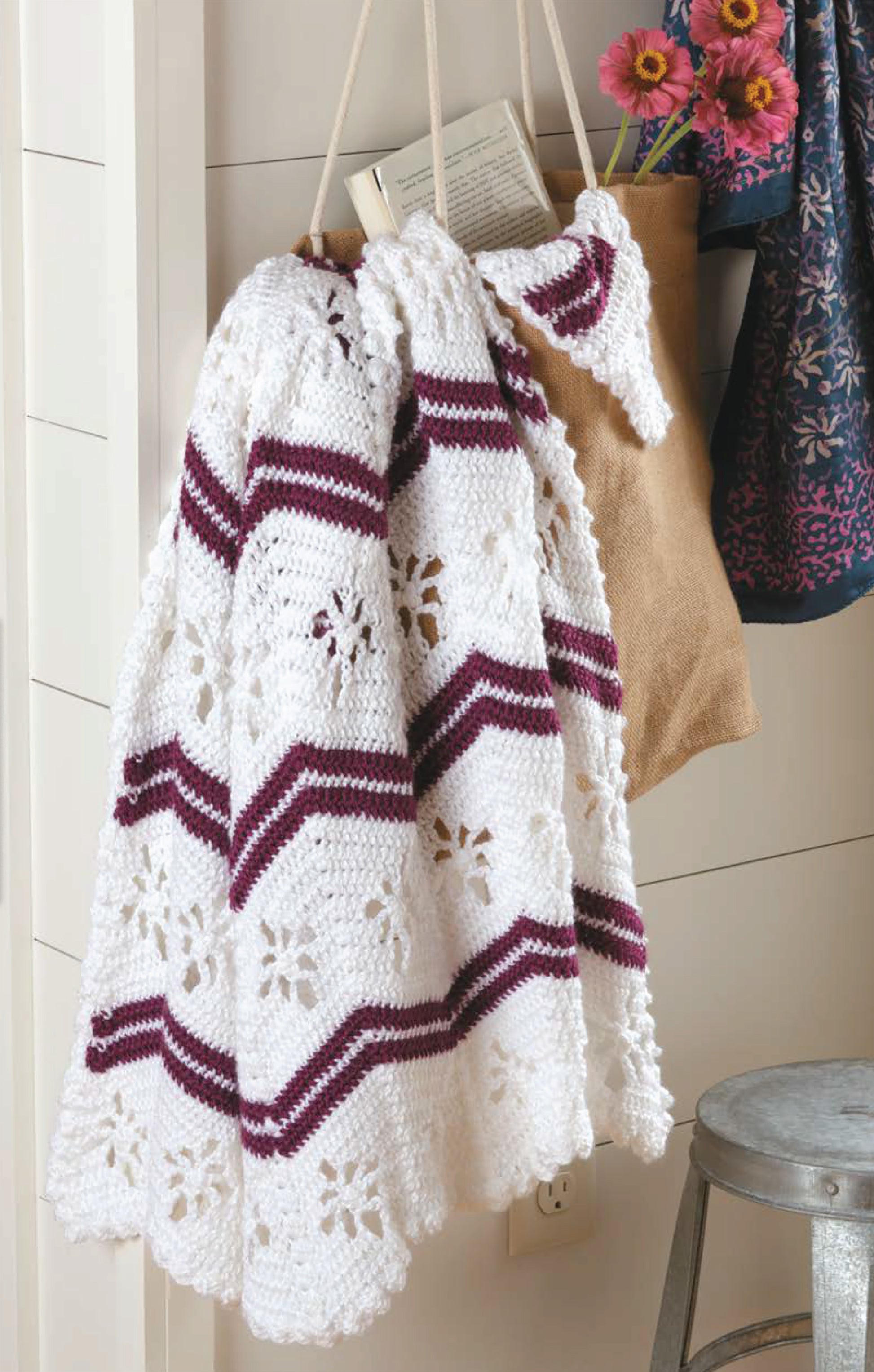 A Chevron Afghans Digital Download by Leisure Arts showcases a white crocheted blanket with maroon and purple chevron stripes and floral patterns hanging beside a burlap tote with pink flowers, a book, and a nearby metal stool.