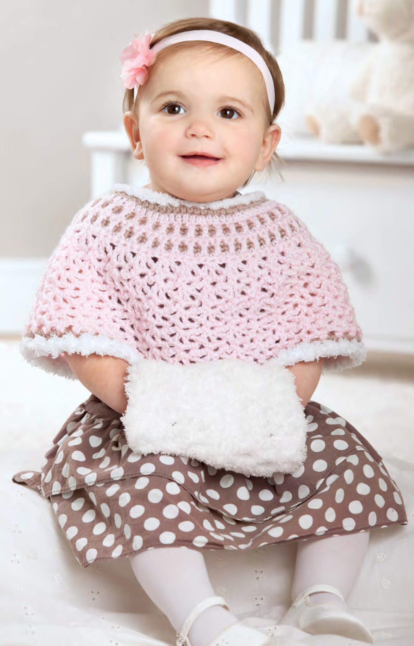 A smiling toddler models a pink crocheted poncho from the Best-Dressed Baby Crochet Digital Download by Leisure Arts, paired with a polka-dot dress and floral headband—showcasing adorable and creative baby designs.