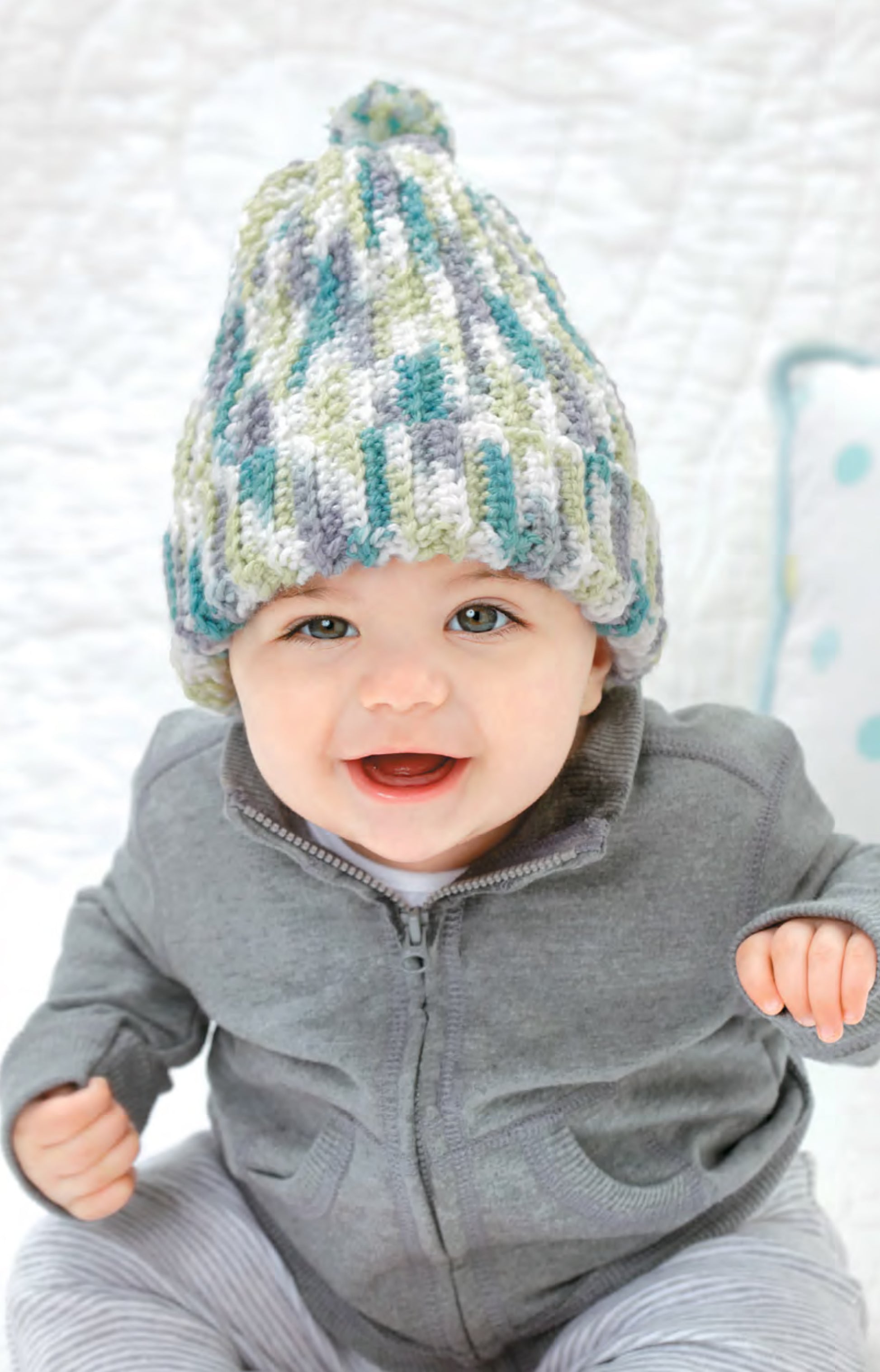 A smiling baby models the Best-Dressed Baby Crochet Digital Download hat by Leisure Arts, featuring a multicolored crochet-inspired design, along with a gray zip-up jacket while sitting on a light surface with a pillow behind.