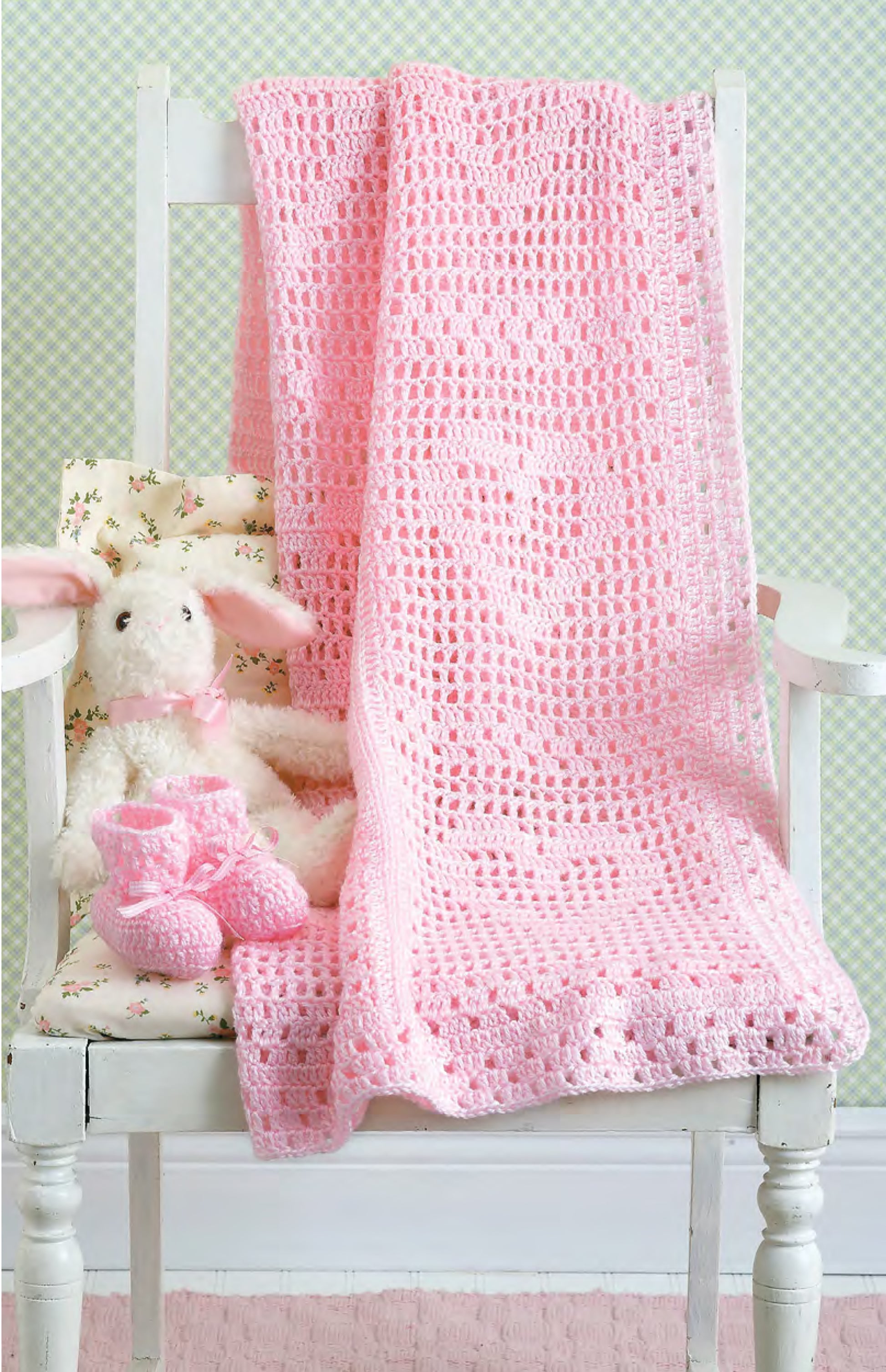 A white wooden chair displays Leisure Arts' Best-Dressed Baby Crochet (digital download), featuring a pink crocheted blanket, a white bunny with pink ears, and pink booties, set against pale green patterned wallpaper.
