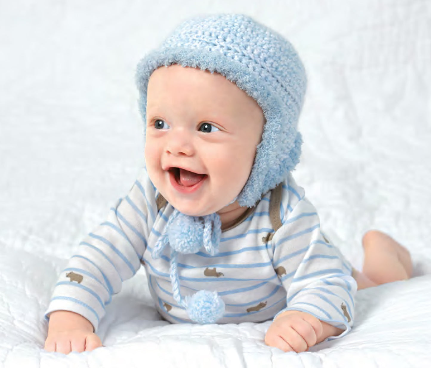 A smiling baby in a blue knit hat with ear flaps and a striped onesie crawls on a white blanket, modeling adorable outfits from the Best-Dressed Baby Crochet Digital Download by Leisure Arts, inspired by cozy crochet designs.