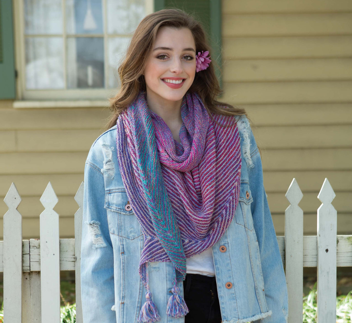 A woman with a pink flower in her hair smiles in her denim jacket and a striped purple scarf. She stands by a white fence, ready to share beginner craft tips using the Boomerang Shawls Digital Download by Leisure Arts.
