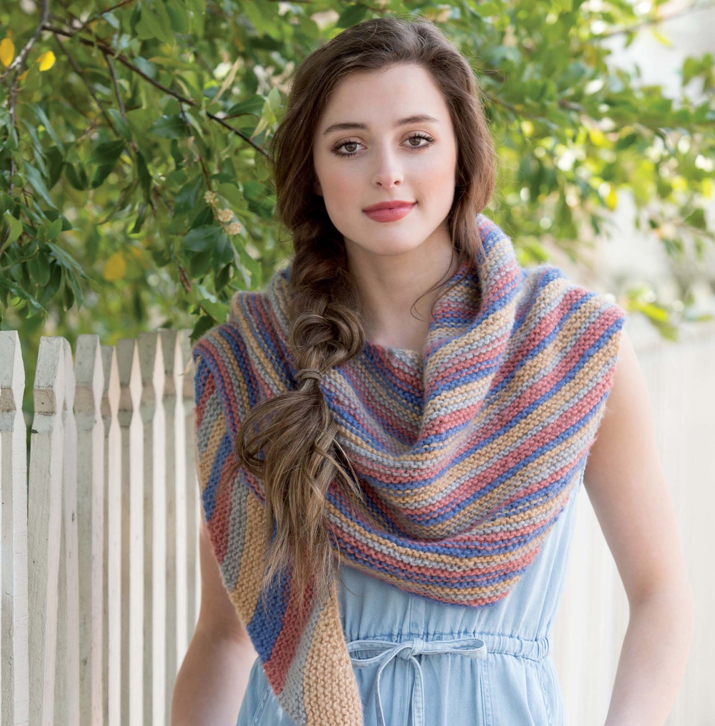 A young woman with long brown hair in a loose braid stands by a white picket fence, modeling the pastel-striped Boomerang Shawls from Leisure Arts—a beginner-friendly digital download—over a light blue dress, with green trees behind her.