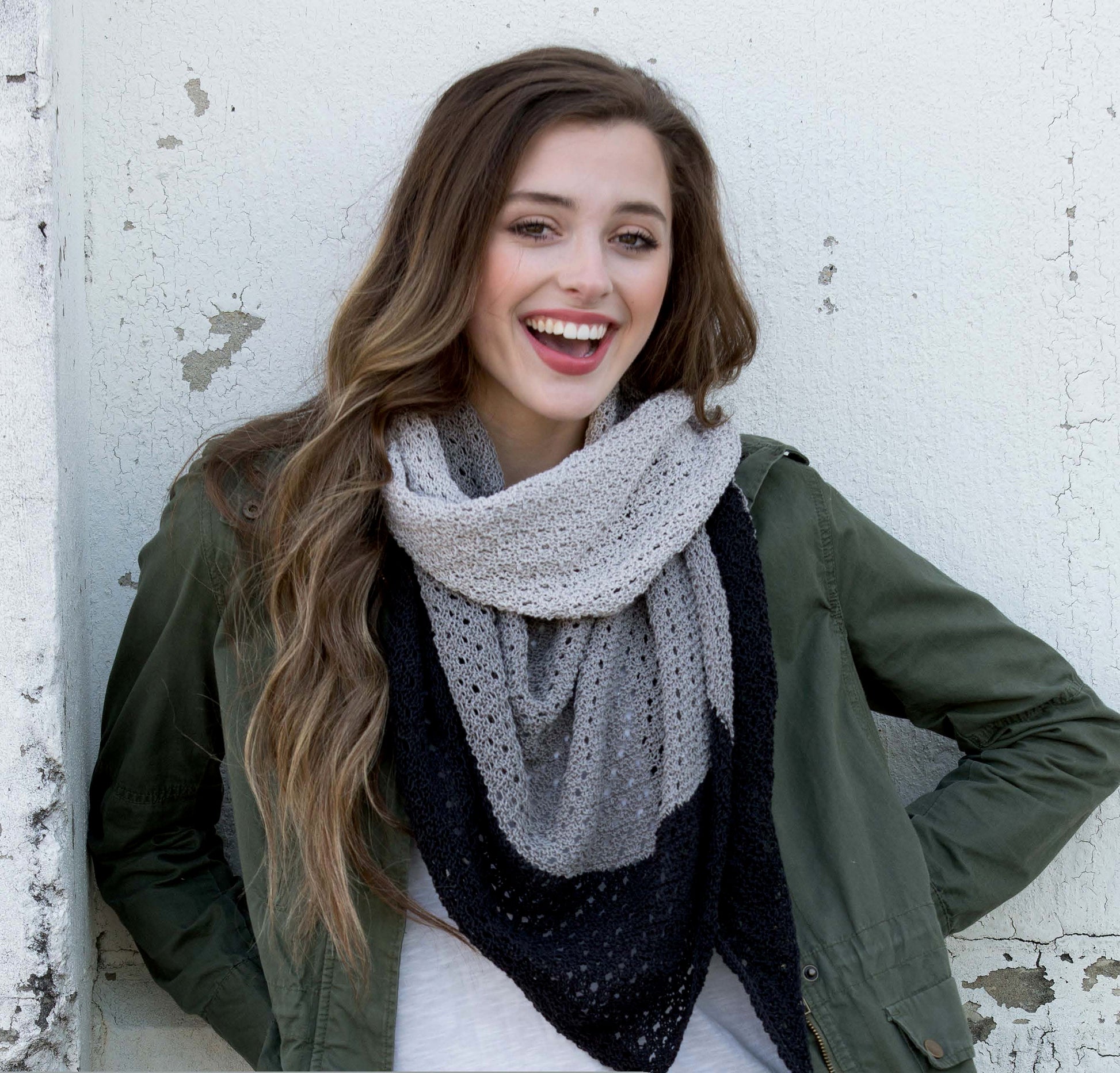 A smiling young woman in a green jacket and knit scarf stands against a textured white wall, ready to share beginner projects using progressive techniques from the Boomerang Shawls Digital Download by Leisure Arts.