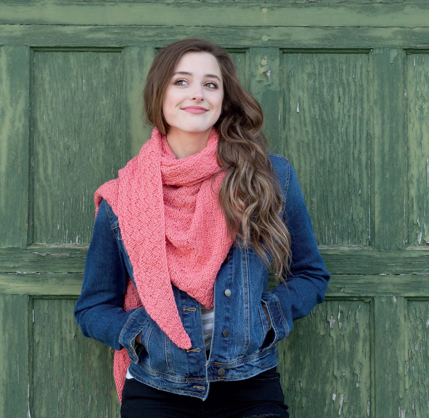A young woman with long wavy hair stands in front of a green wooden door, wearing a denim jacket and a pink shawl she made from the Boomerang Shawls Digital Download by Leisure Arts, her hands in her pockets and a slight smile on her face.