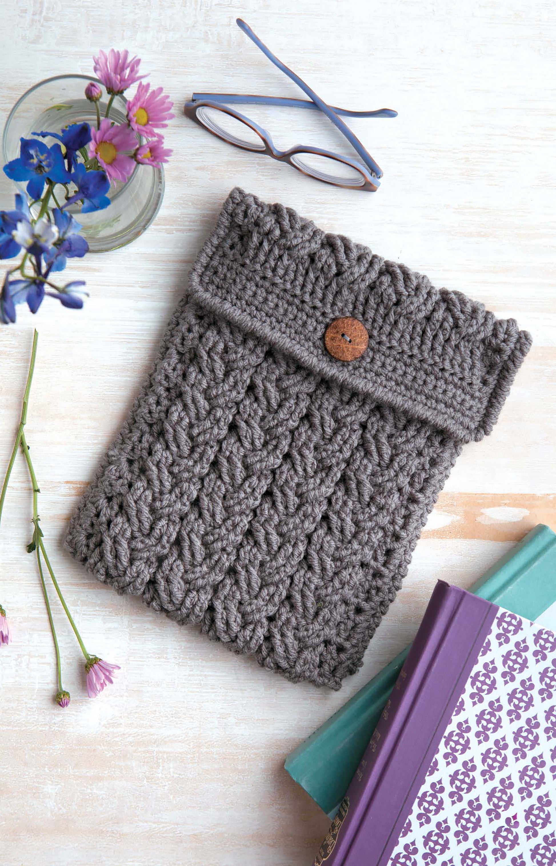 A gray crocheted pouch made with worsted weight yarn and a wooden button sits beside a vase, eyeglasses, and books—ideal inspiration from Leisure Arts’ Crochet Bags & Backpacks for crafters seeking new patterns.