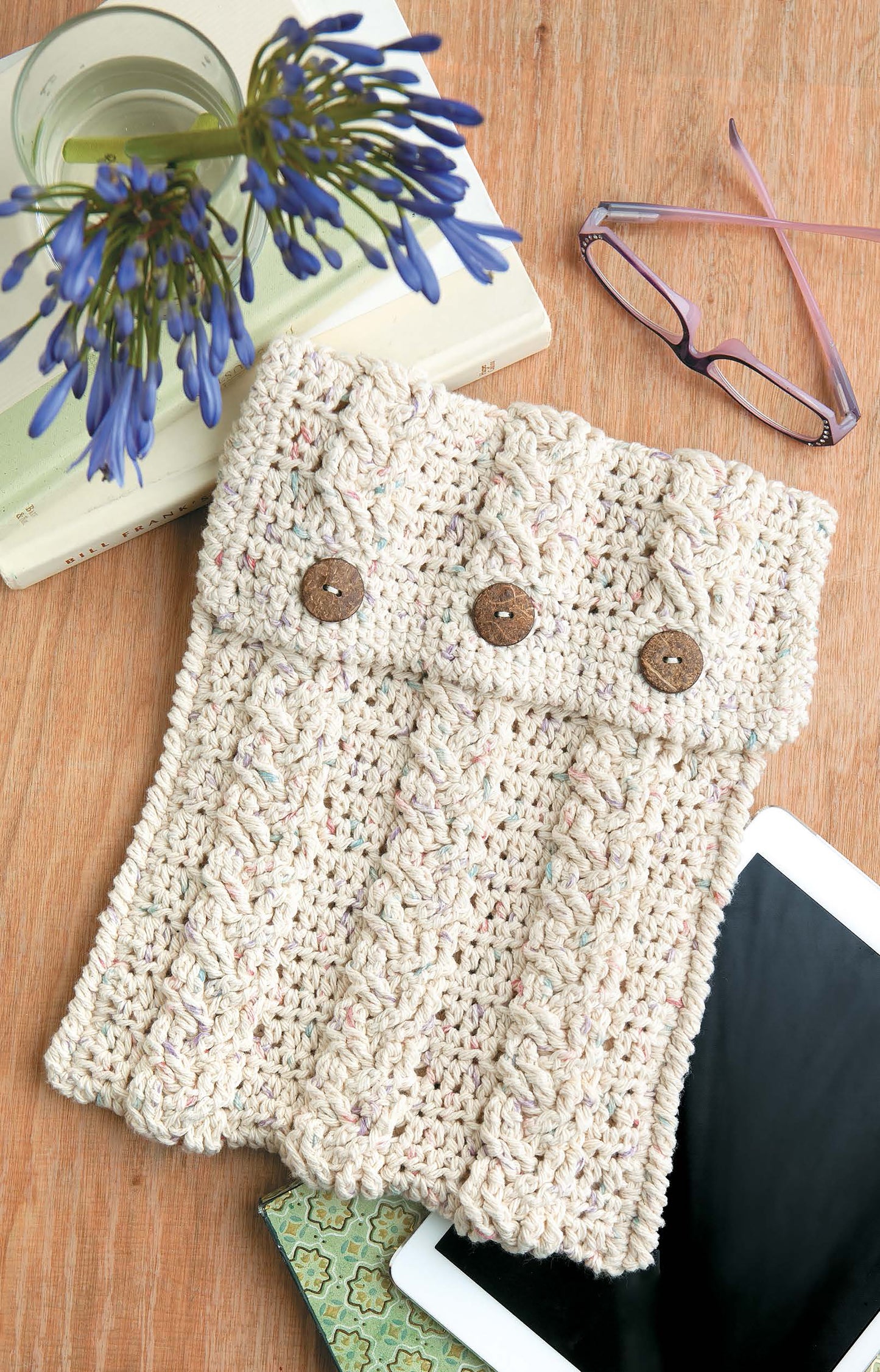 The Leisure Arts Crochet Bags & Backpacks beige tablet sleeve, crocheted with worsted weight yarn and three brown buttons, rests on a wooden surface—an ideal intermediate project for stylish tech protection.