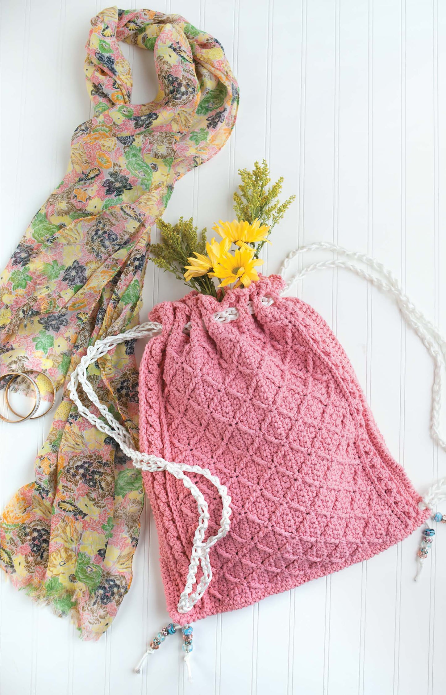 The Crochet Bags & Backpacks by Leisure Arts features a pink crocheted drawstring bag with white braided handles, shown holding yellow flowers next to a floral scarf and gold bangles on a white surface.