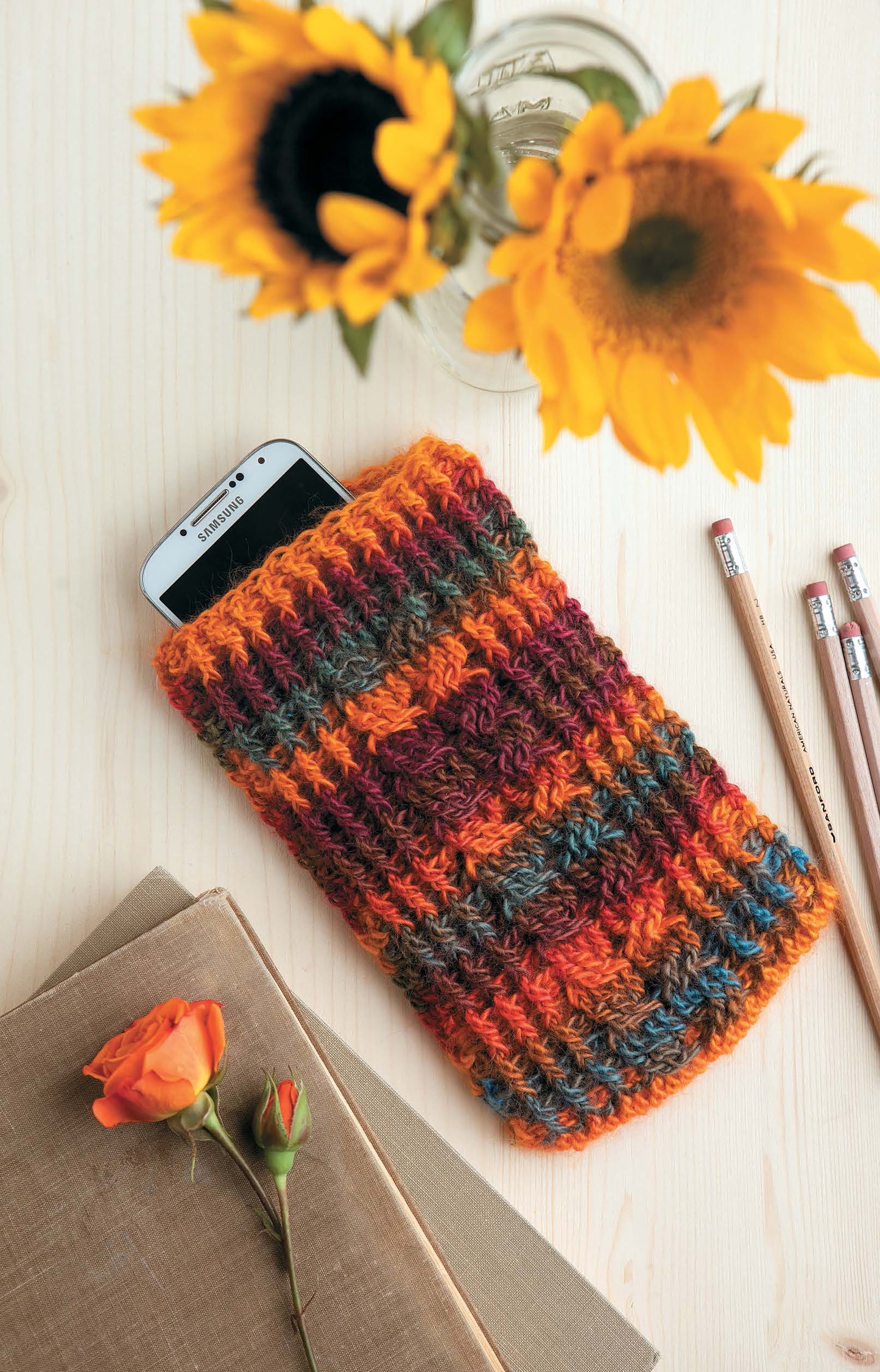 A white smartphone in a colorful case made with worsted weight yarn from Leisure Arts’ Crochet Bags & Backpacks rests on a light wooden table beside sunflowers in a glass jar, two pencils, a closed book, and an orange rose.