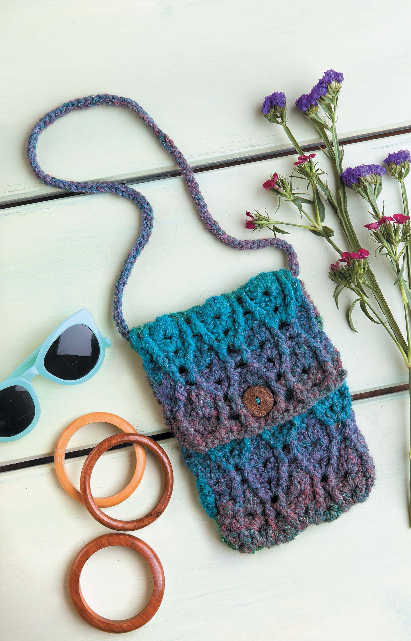 The Crochet Bags & Backpacks by Leisure Arts, crafted with worsted weight yarn in blue and purple and finished with a button closure, rests on a light surface beside sunglasses, wooden bangles, and purple flowers—ideal for intermediate crocheters.