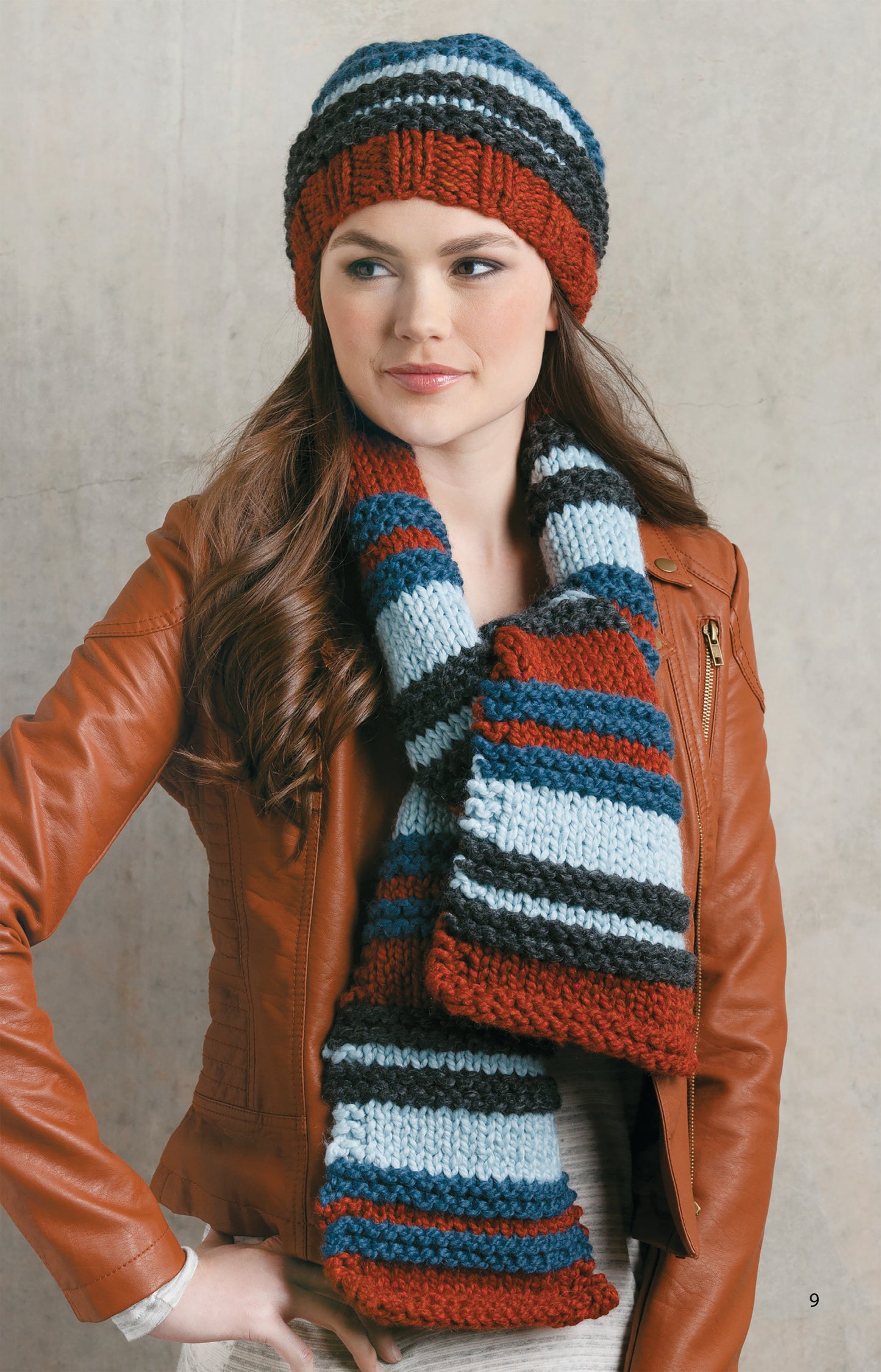 A woman wears a brown leather jacket, matching scarf, and a multicolored striped knit hat from Chunky Winter Knits by Leisure Arts—ideal for beginner knitters—while standing against a neutral background.