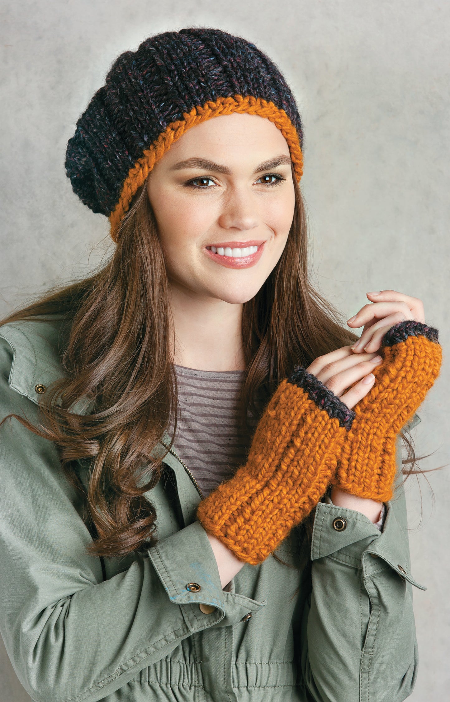 A woman models the Chunky Winter Knits hat and fingerless gloves by Leisure Arts—super bulky yarn in orange and dark purple—paired with a green jacket and striped shirt, set against a neutral background.