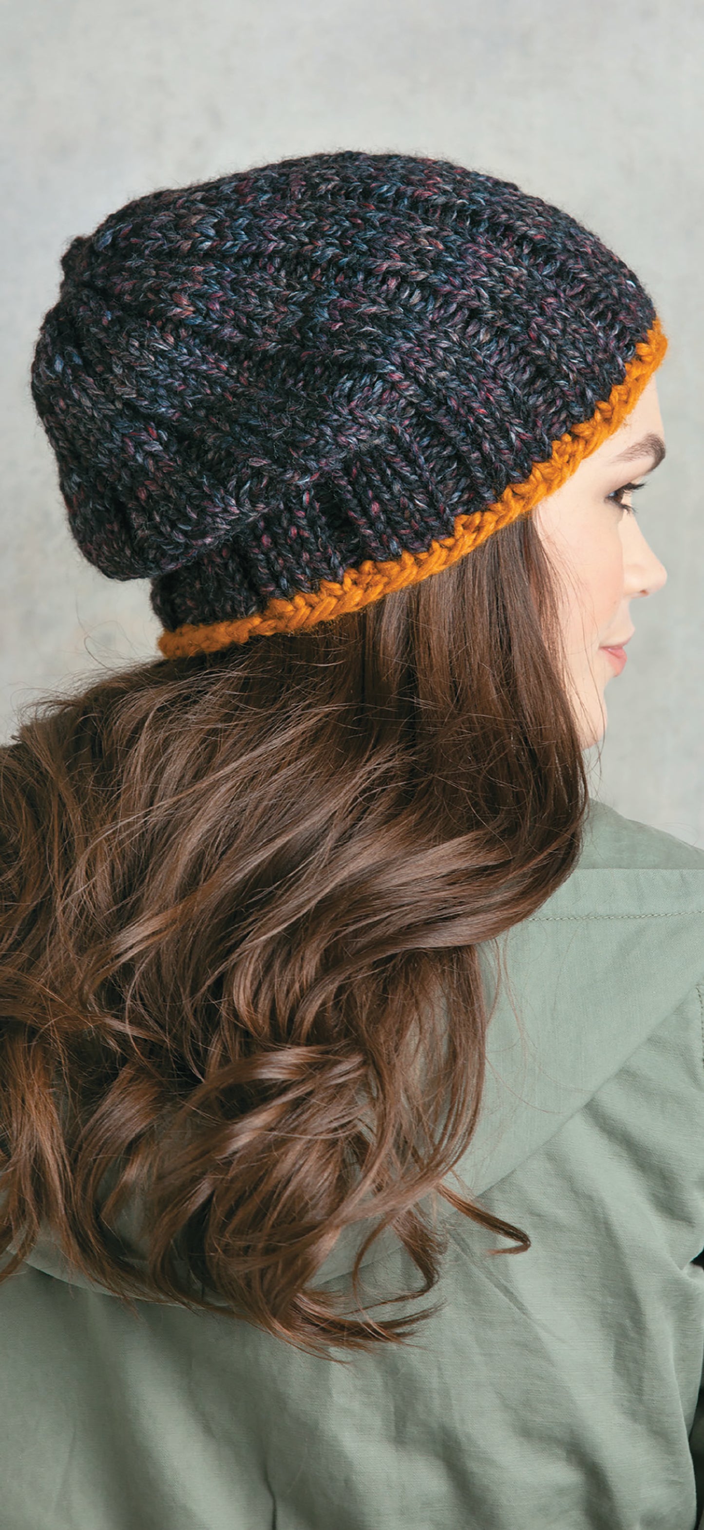A woman with long, wavy brown hair is shown from behind wearing a dark knit beanie with an orange band made from Chunky Winter Knits by Leisure Arts. She wears a green jacket and faces right against a neutral background.