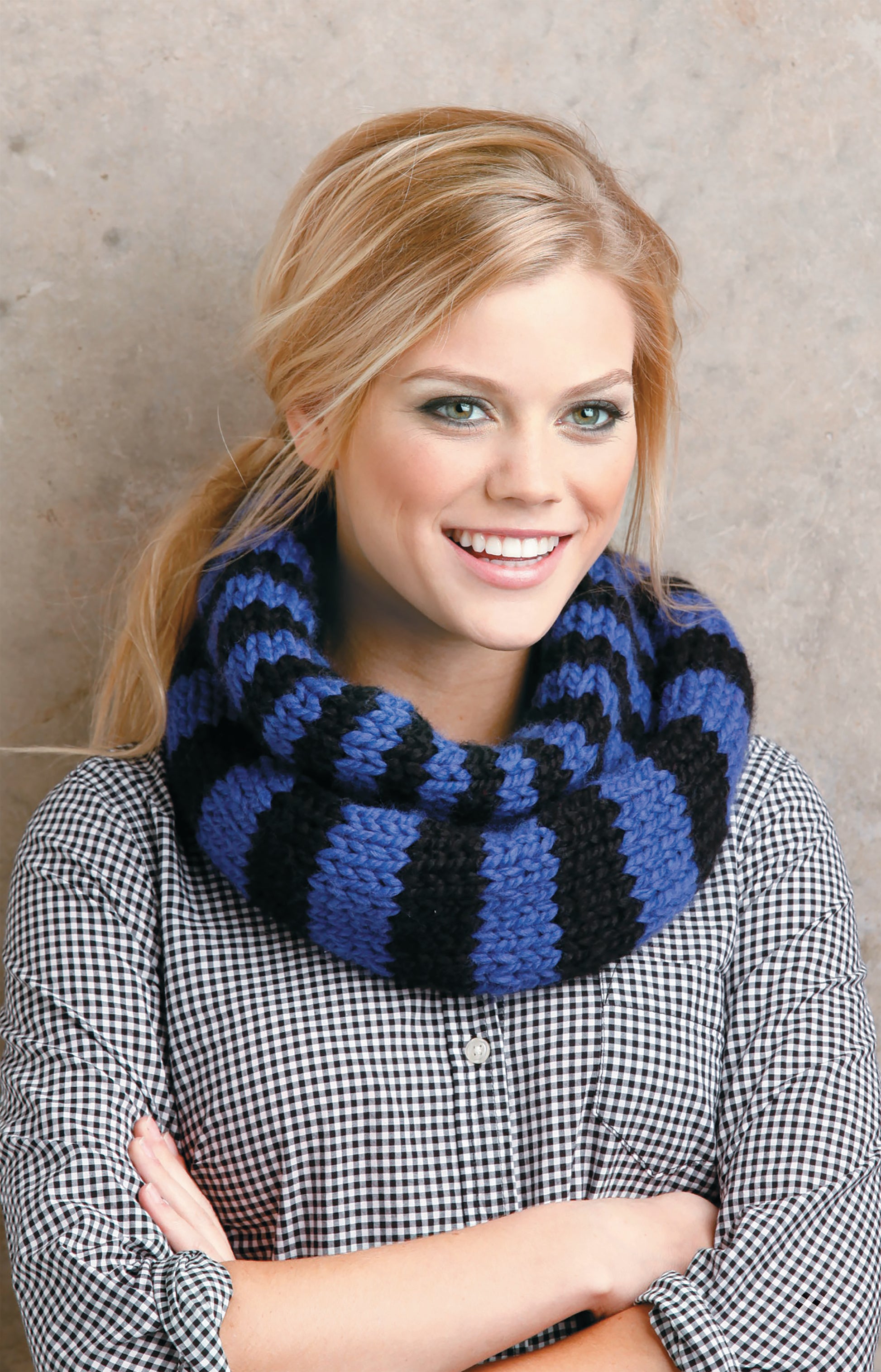A smiling blond woman models the Chunky Winter Knits scarf by Leisure Arts—a black and blue striped, super bulky knit ideal for beginners—paired with a black-and-white checkered shirt against a textured beige wall.