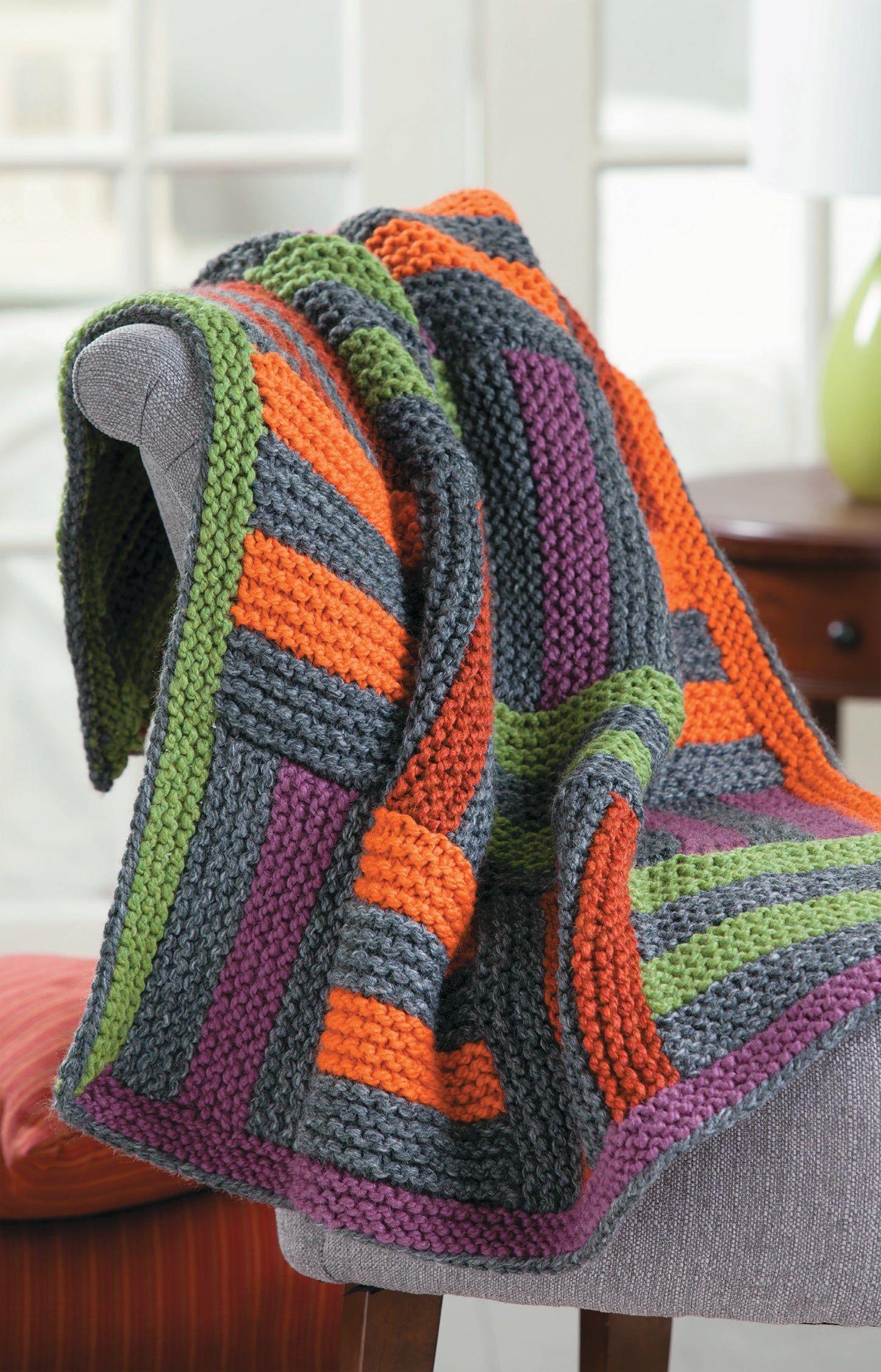 Chunky Winter Knits by Leisure Arts features a super bulky, multicolored striped blanket (gray, orange, green, purple) draped over a gray chair—ideal for beginner knitting projects in bright rooms.
