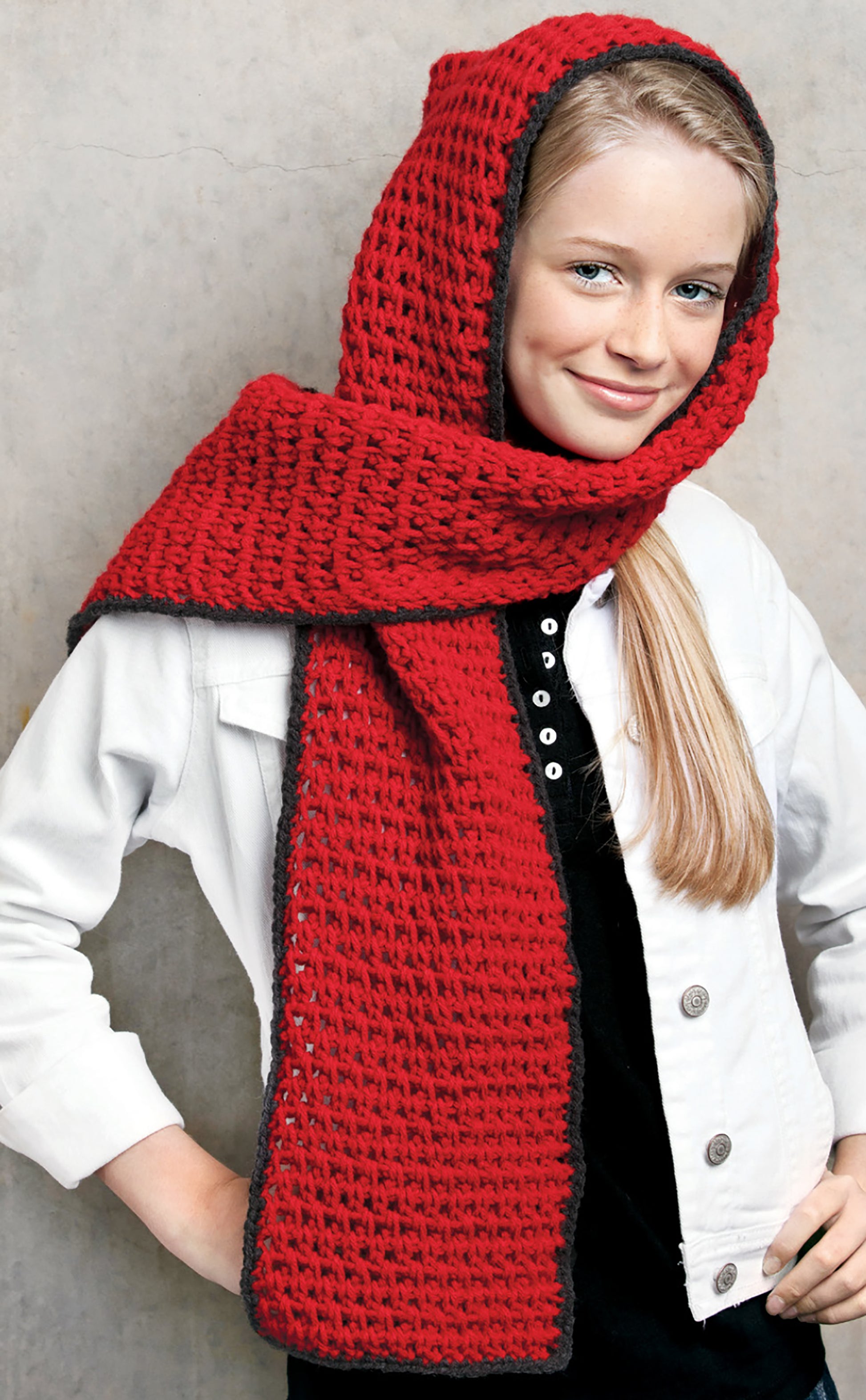 A young person with long blonde hair smiles in a white jacket and black top, modeling a bright red hooded scarf from Crochet Family Favorites by Leisure Arts, shown against a plain gray background.