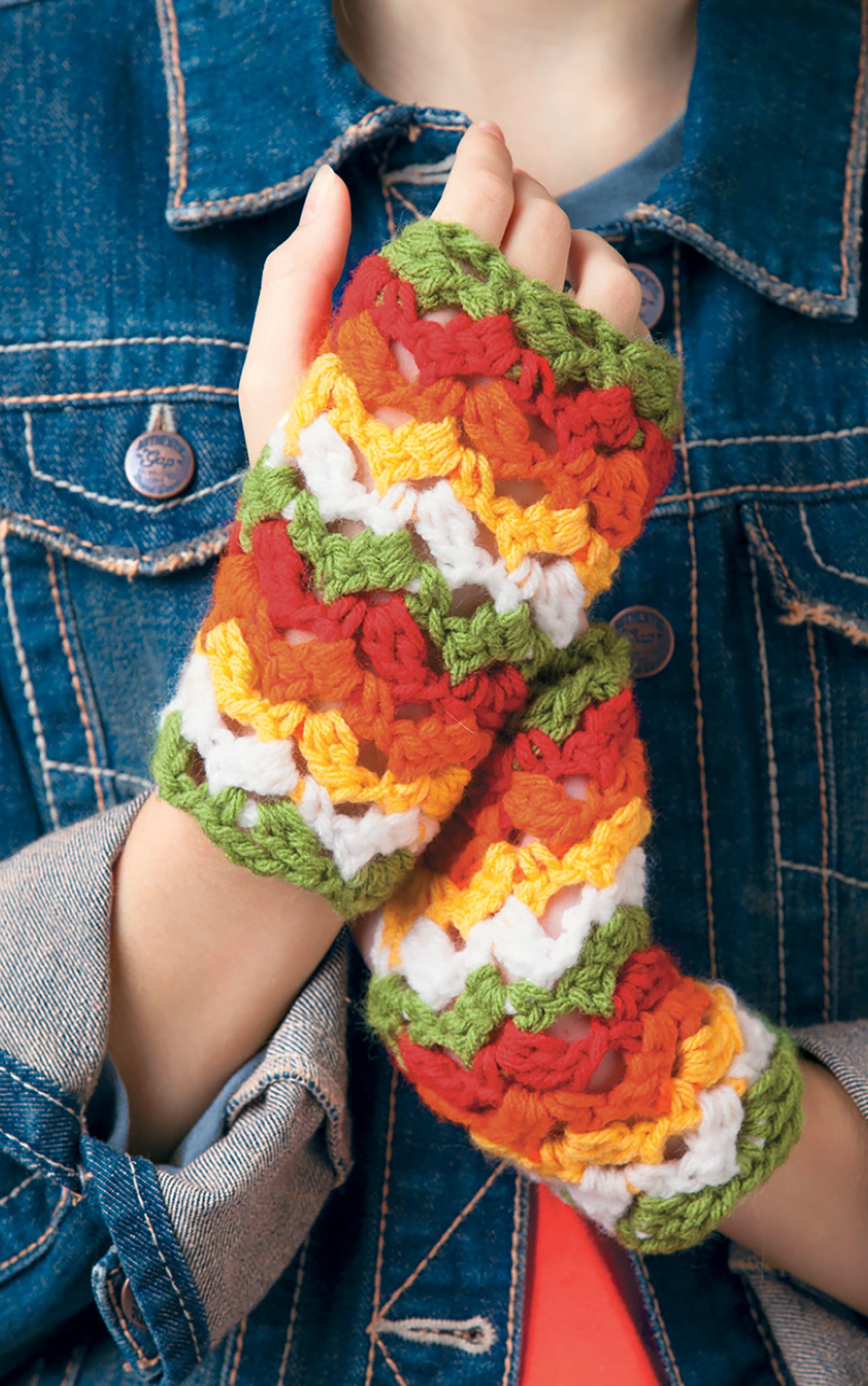 A model in a denim jacket showcases Leisure Arts' Crochet Family Favorites: vibrant, scalloped fingerless gloves in red, orange, yellow, green, and white stripes. The model’s nails are unpainted.