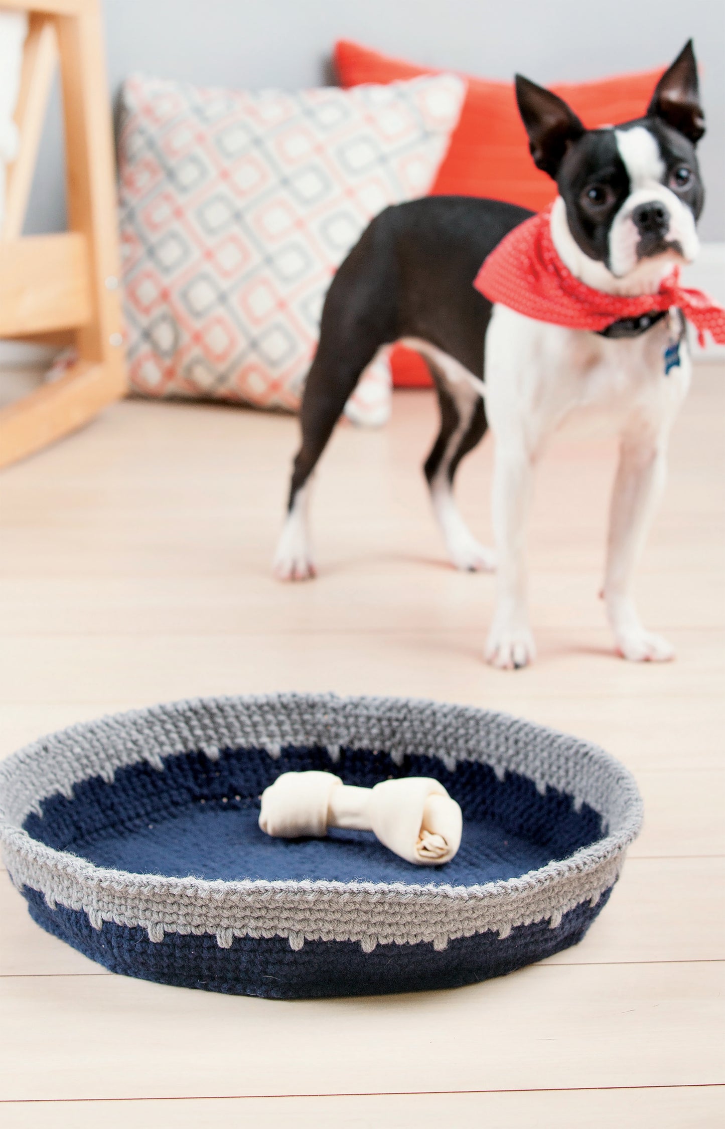 A Boston Terrier in a red bandana stands on a wooden floor behind the Leisure Arts Crochet Family Favorites dog bed, featuring blue and gray yarn with a rawhide bone inside. Patterned and plain pillows are in the background.